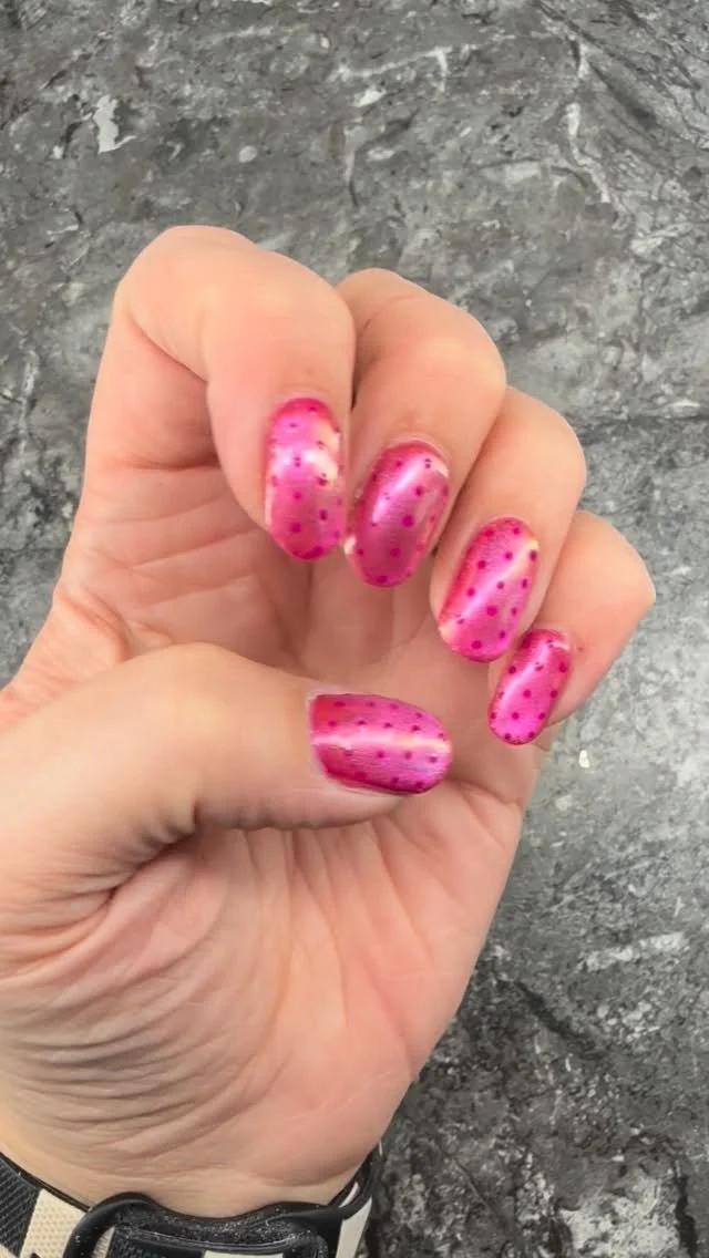 Close-up of a hand with pink nail polish and red polka dots, set against a gray textured background.