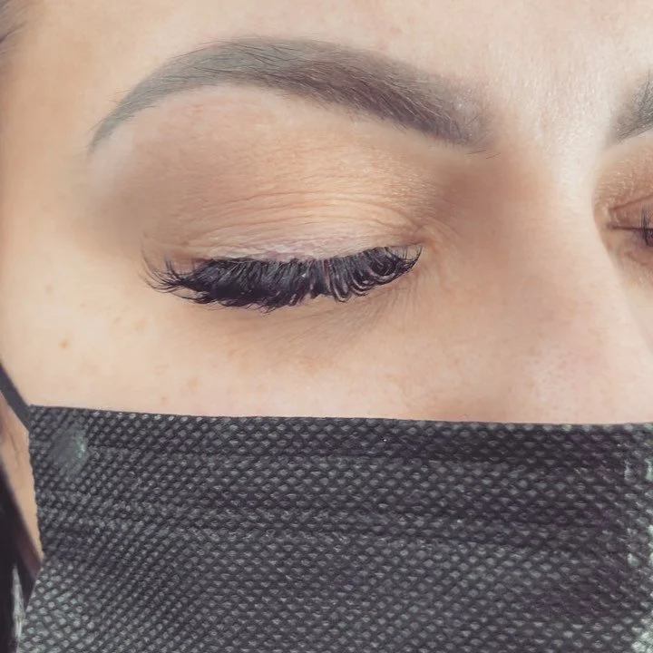 Close-up of a person's eye with makeup, closed eyelid, and long curled eyelashes, partially covered by a black face mask.