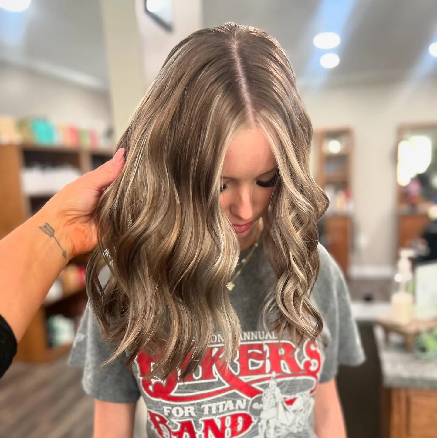 A woman with long, wavy blonde hair looking down as a person touches her hair. She is wearing a gray T-shirt with red and white text and graphics, and there are shelves and light fixtures in the background.