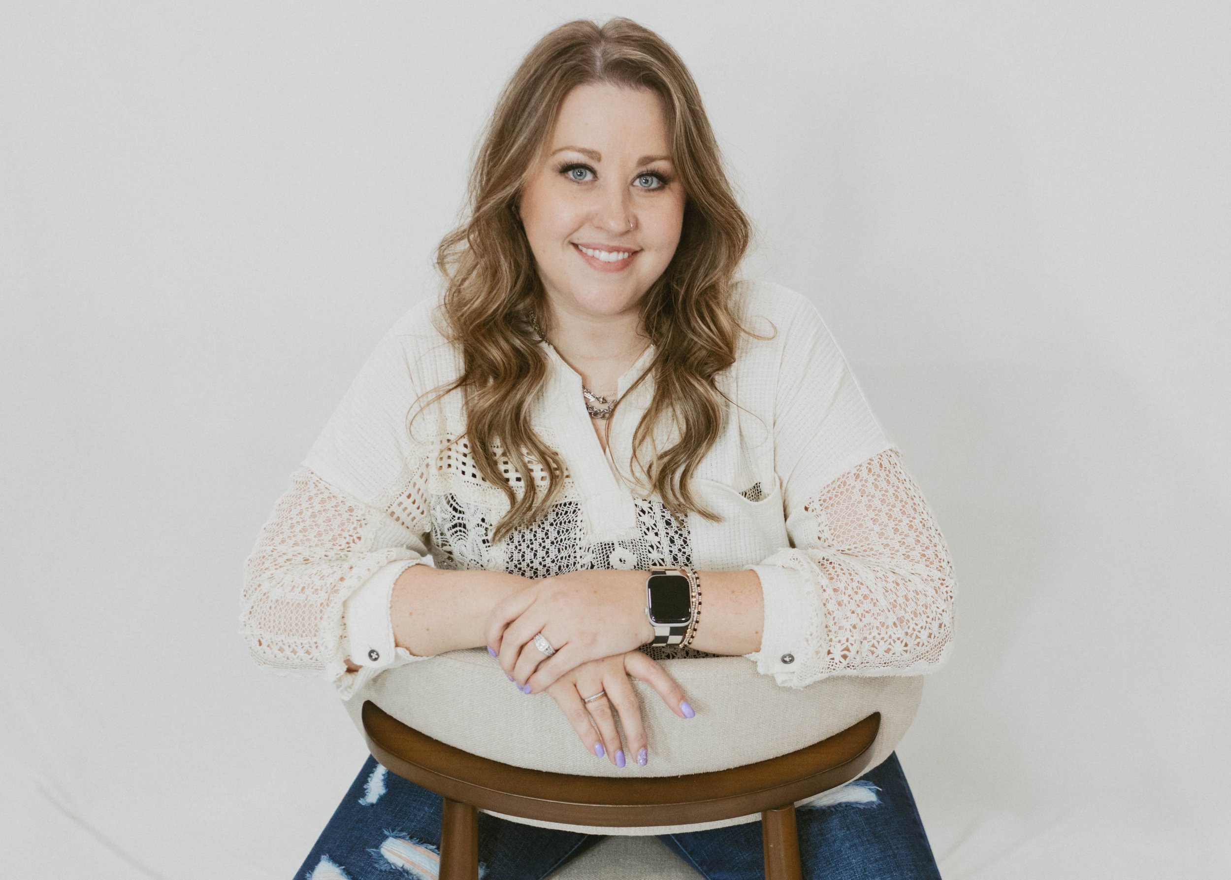 Smiling woman with wavy, shoulder-length hair sitting behind a chair with crossed arms, wearing a cream lace top, blue jeans, and a smartwatch.