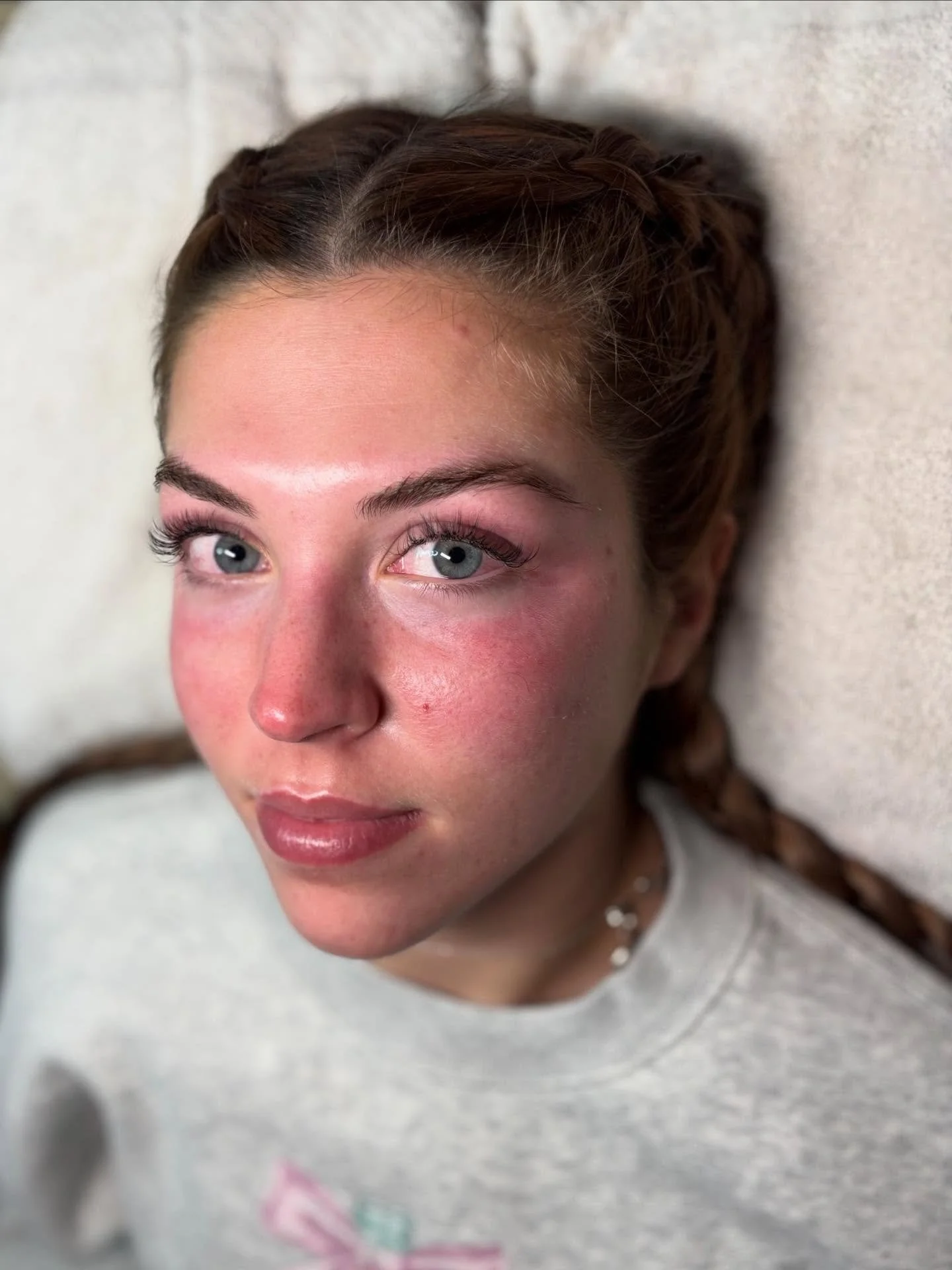 A young woman with braided hair, blue eyes, and a slight smile, lying on a light-colored surface, wearing a gray shirt with a small design and a choker necklace.