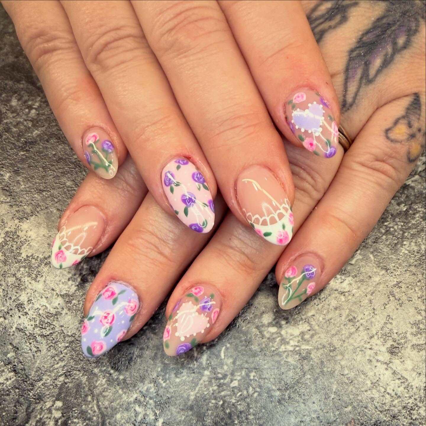 Nail art on fingernails with pastel floral designs and lace patterns on a beige and purple background.