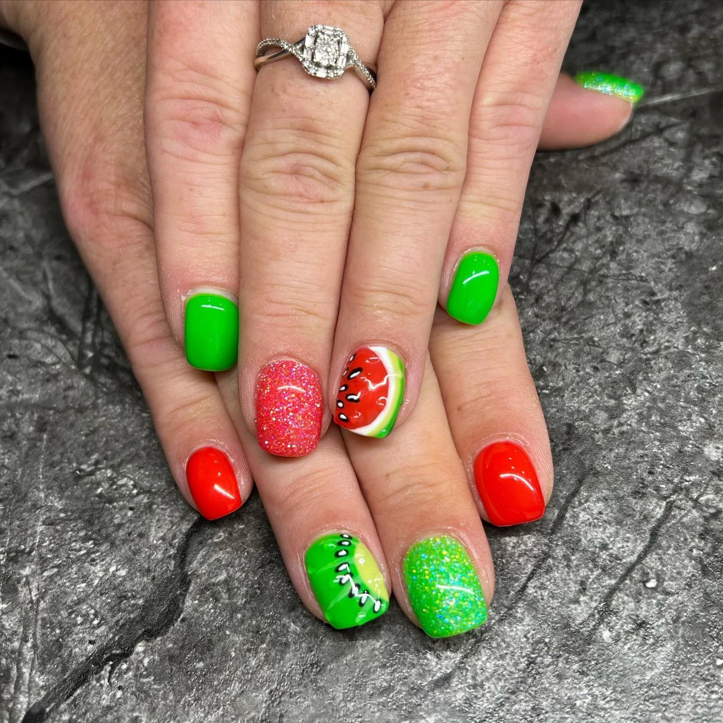 Colorful nail art with watermelon and strawberry themes on fingernails, green, red, and glitter pink polish, with a silver engagement ring with a large rectangular diamond on the finger.