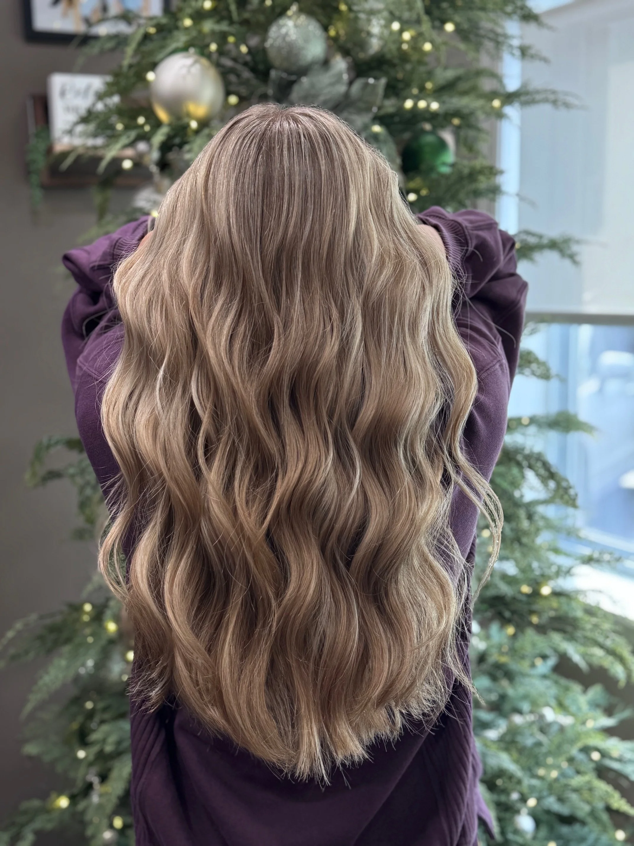 Back of a woman with long, wavy blonde hair standing in front of a decorated Christmas tree.