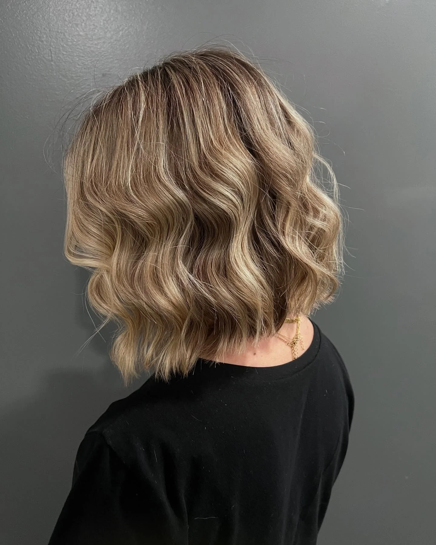 Side view of a woman with shoulder-length blonde wavy hair, wearing a black shirt and a gold necklace against a gray background.