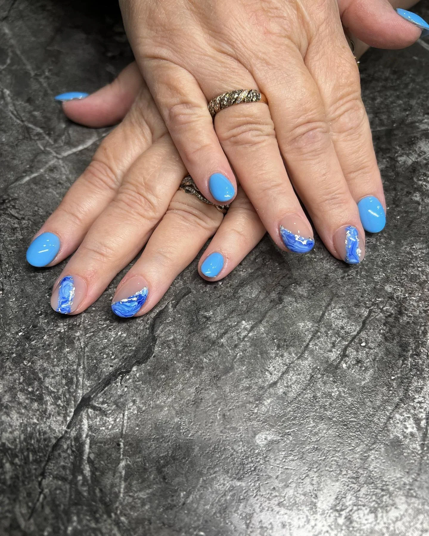 Close-up of hands with manicured nails, featuring blue polish and marble design on the ring and middle fingers, resting on a textured dark surface. The person is wearing two rings on the middle finger.