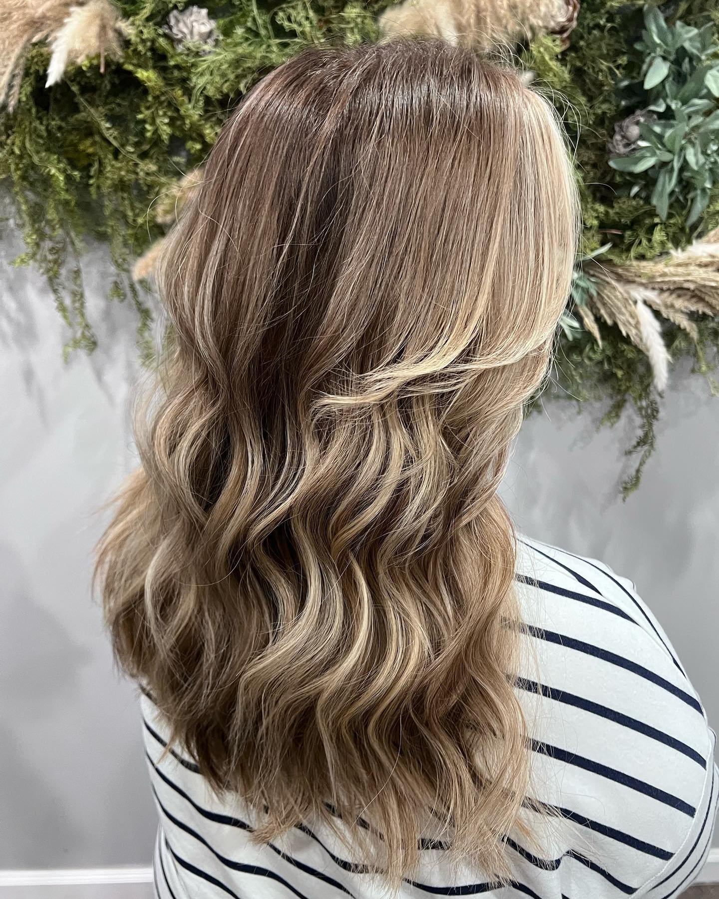 Back view of a woman with long, wavy, blonde hair standing in front of a green, leafy background.
