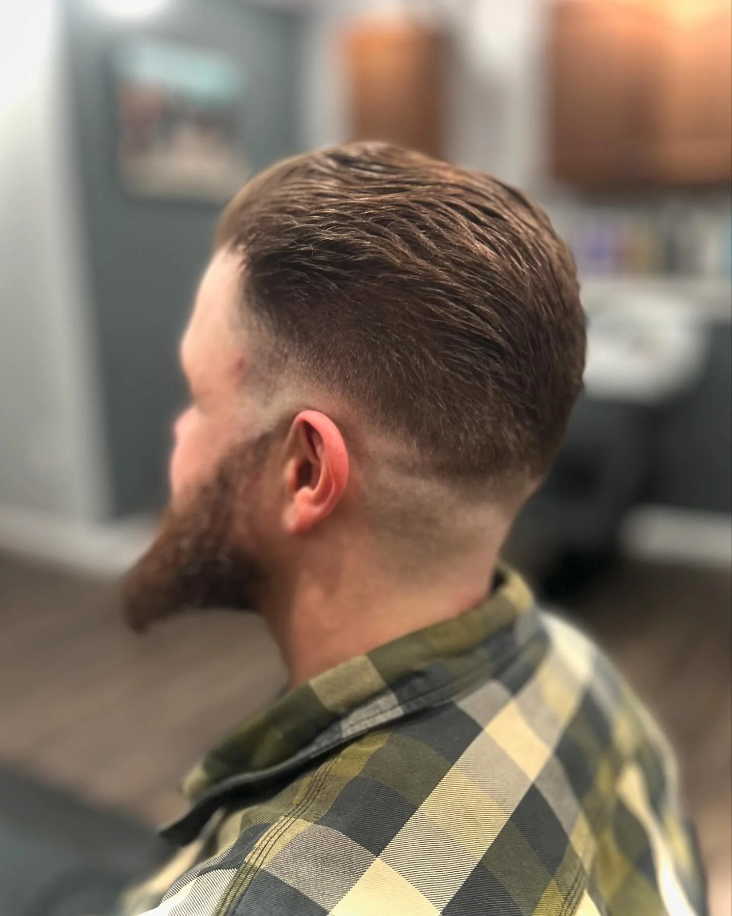 Side view of a man with a fresh fade haircut and a beard, wearing a plaid shirt, in a barbershop or salon.