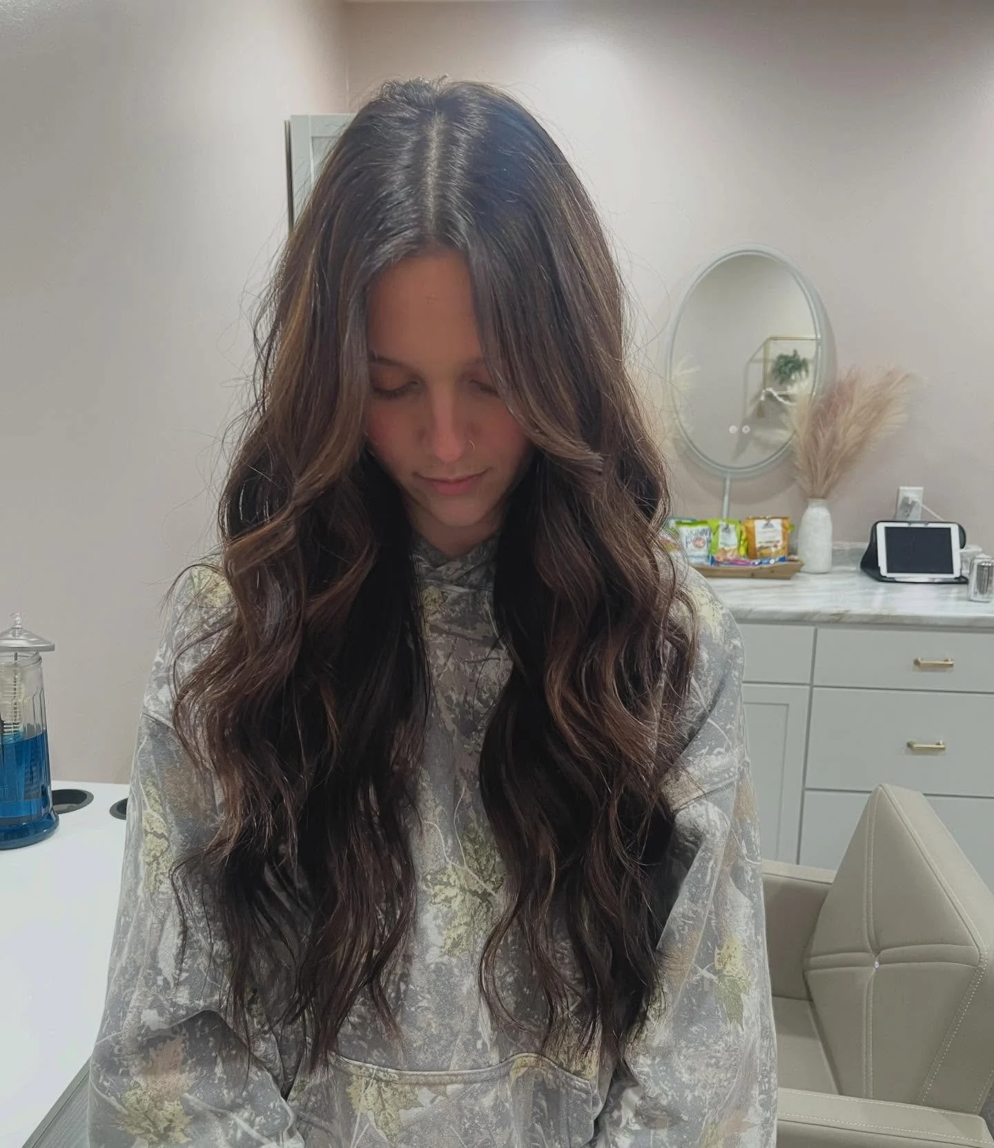 A woman with long, wavy dark hair looking down in a room with light-colored walls and furniture. There are health vials, a mirror, and a tablet on a white cabinet in the background.
