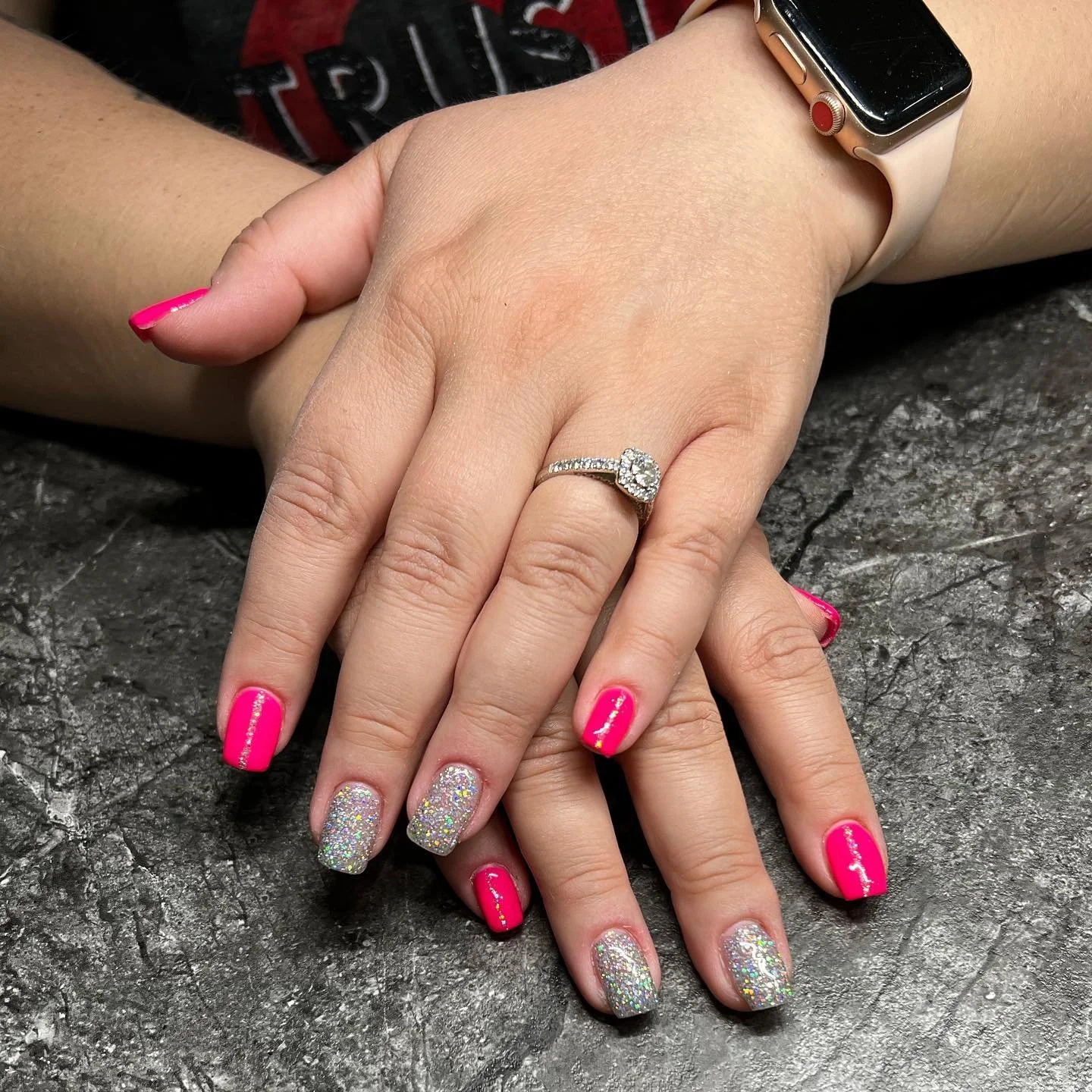 Close-up of two hands with pink and glitter nail polish, displaying an engagement ring with a large center diamond and smaller diamonds on a silver band, resting on a dark stone surface. One hand has a smartwatch with a pink band.