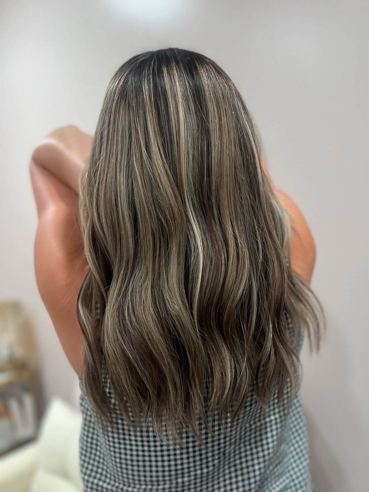 Back view of a woman with long wavy hair that is brown with blonde highlights, wearing a checkered shirt.