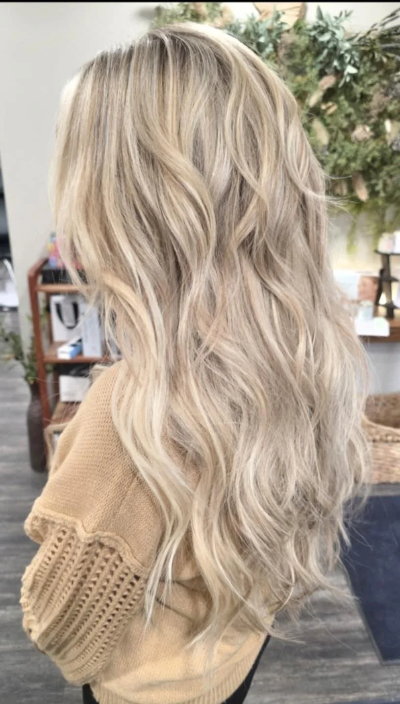 Side view of a woman with long, wavy, blonde hair wearing a beige sweater in a room with plants and shelves.