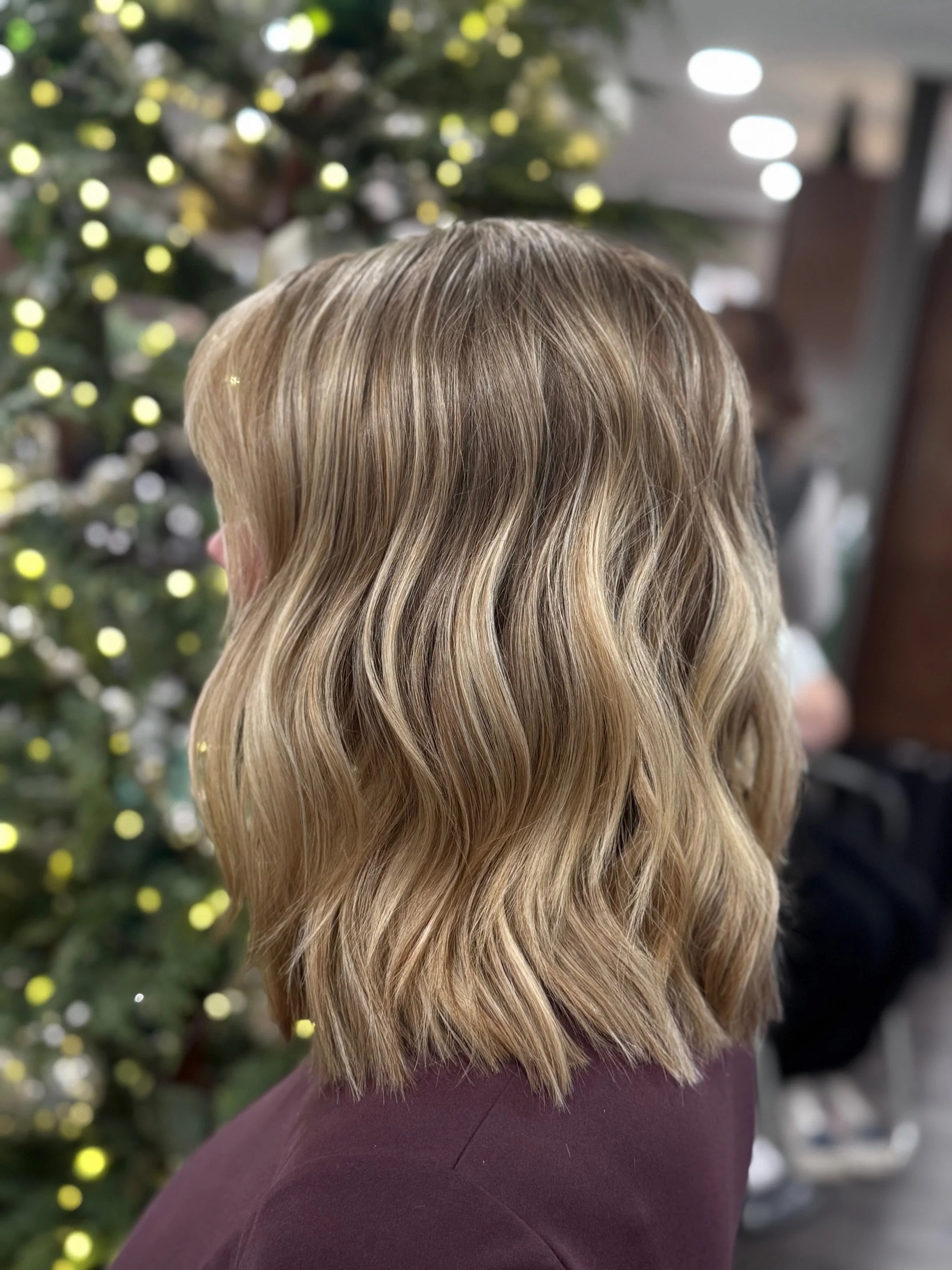 Close-up of a woman with shoulder-length wavy blonde hair, standing in front of a decorated Christmas tree with yellow and white lights.