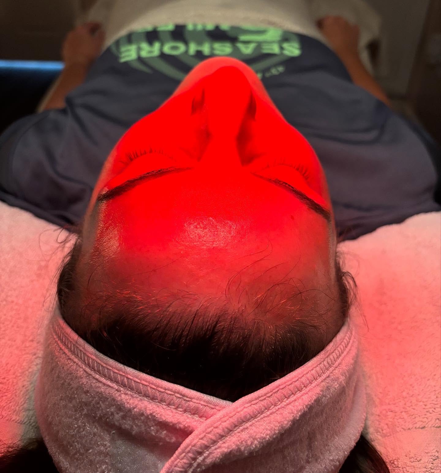 A person lying down with their eyes closed, receiving a red LED light therapy treatment on their face, wearing a headband and casual clothing.