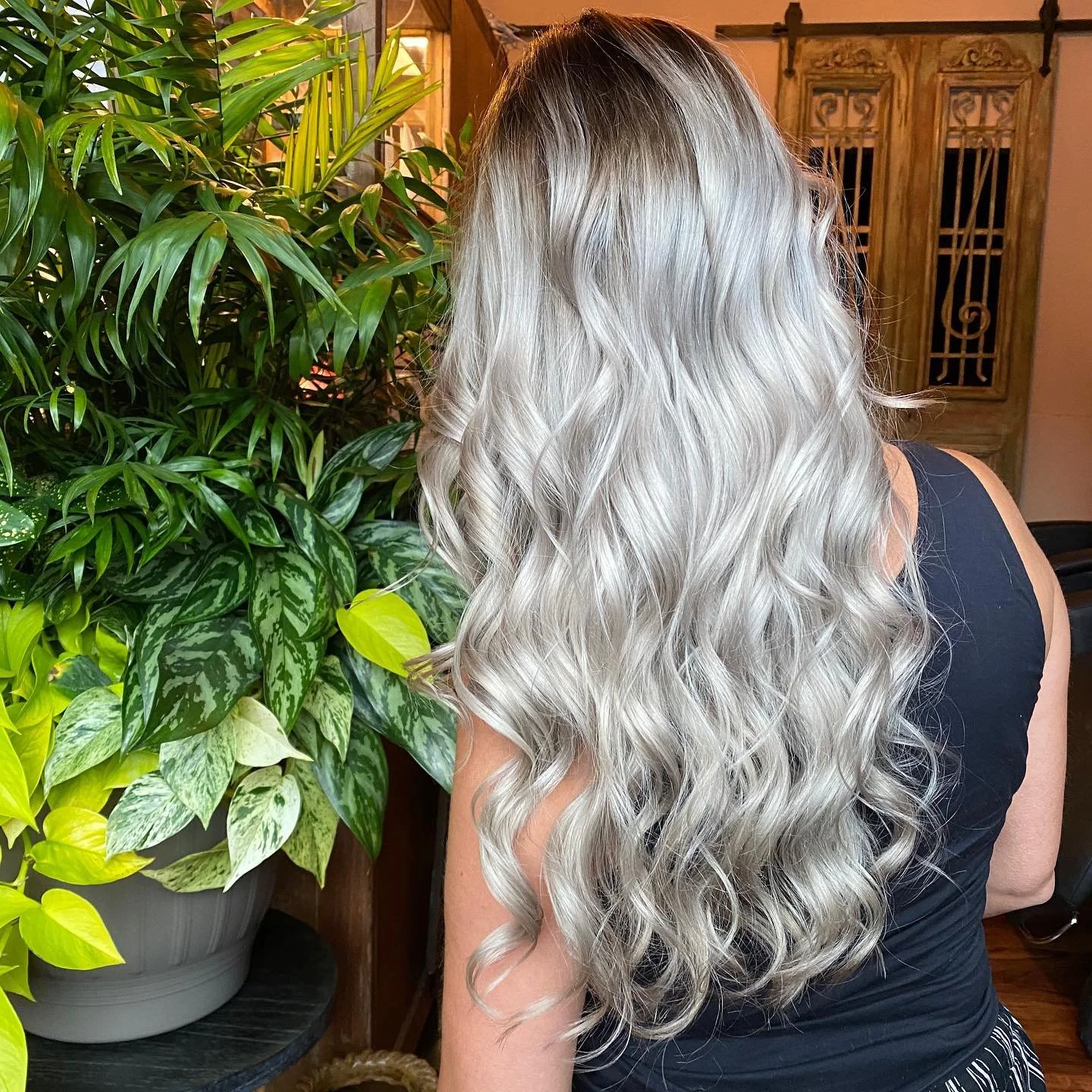 A woman with long, blonde, curly hair standing near green houseplants.
