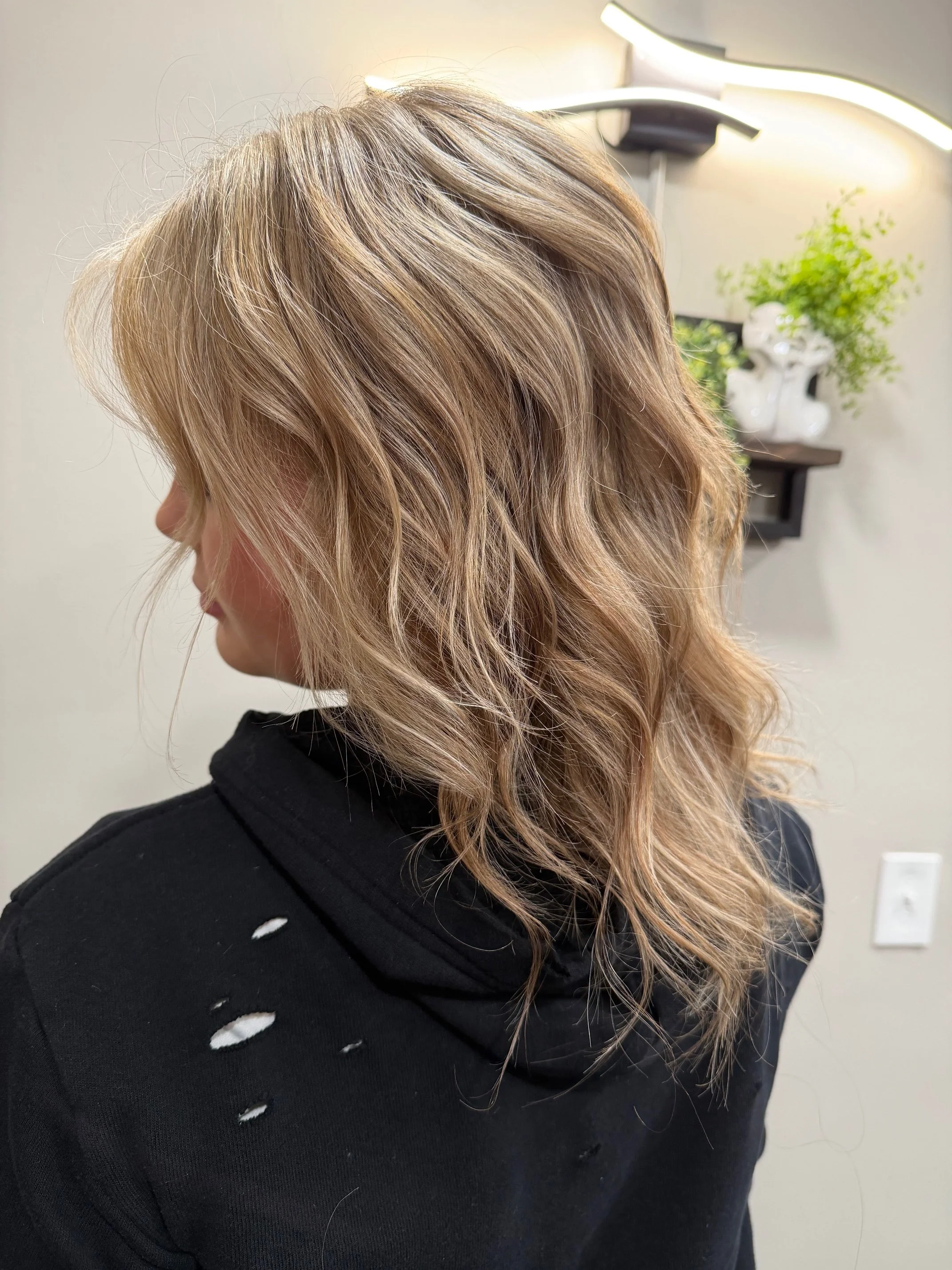 Side view of a woman with shoulder-length, wavy blonde hair with darker roots, wearing a black distressed shirt.