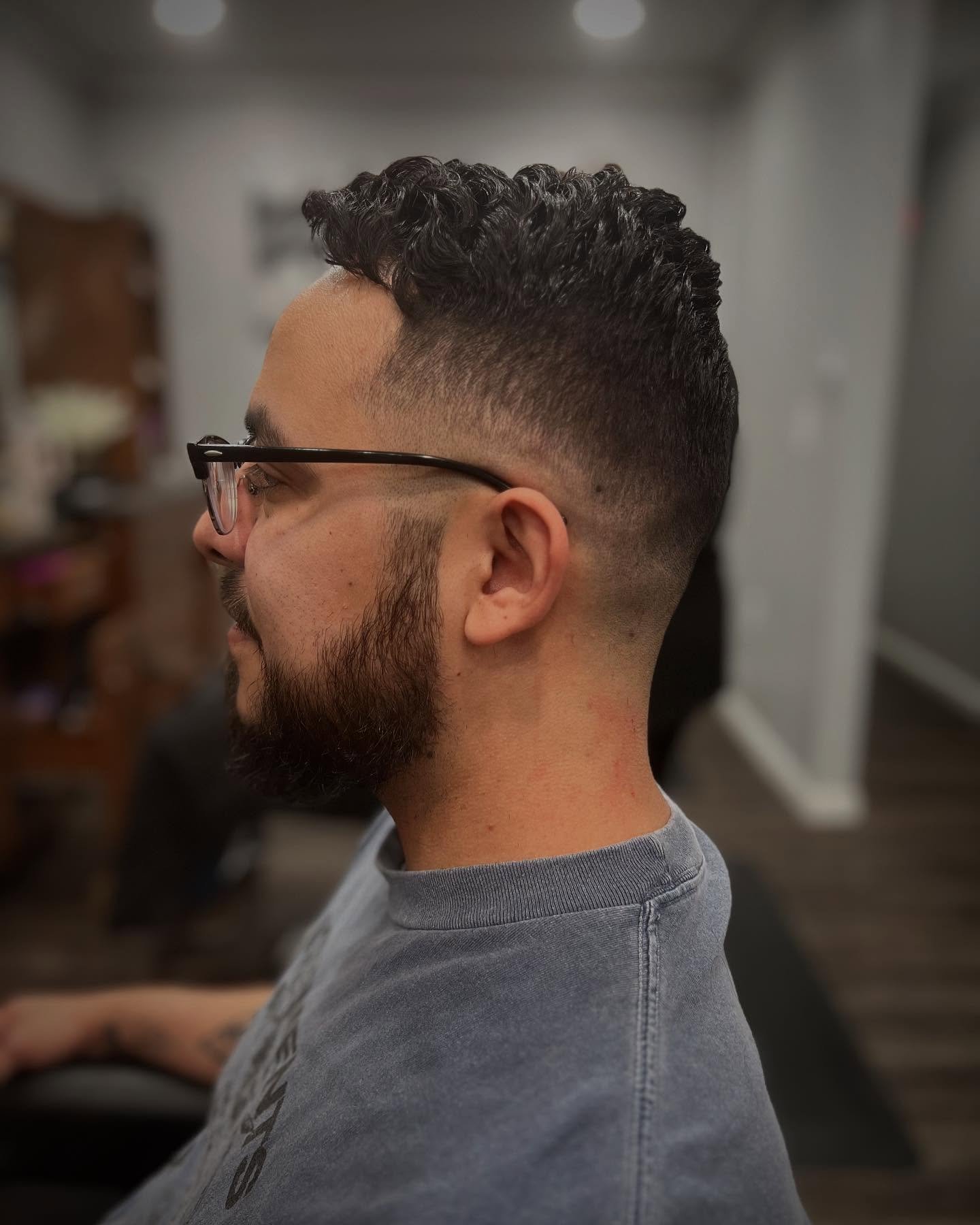 Side view of a man with a modern fade haircut and beard, wearing glasses and a gray T-shirt, sitting in a barbershop.