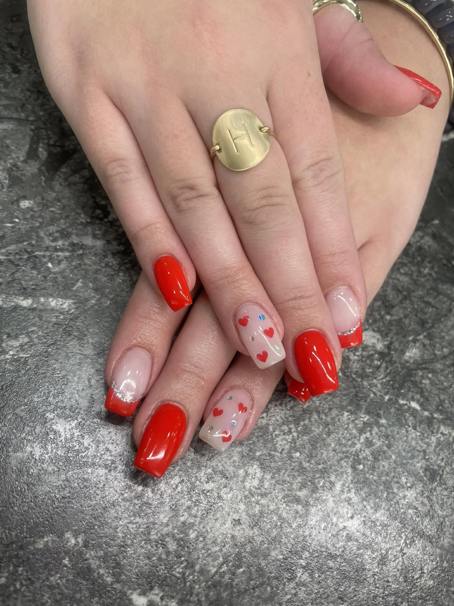 Close-up of hands with decorated nails and jewelry, one hand resting on the other on a textured gray surface.