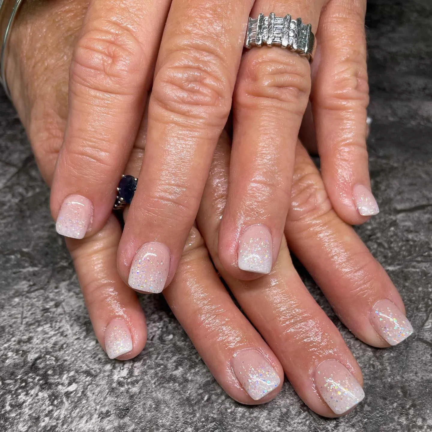 Hands with well-manicured nails featuring white glitter nail polish; one hand has a silver ring with a rectangular design; the background is a dark textured surface.