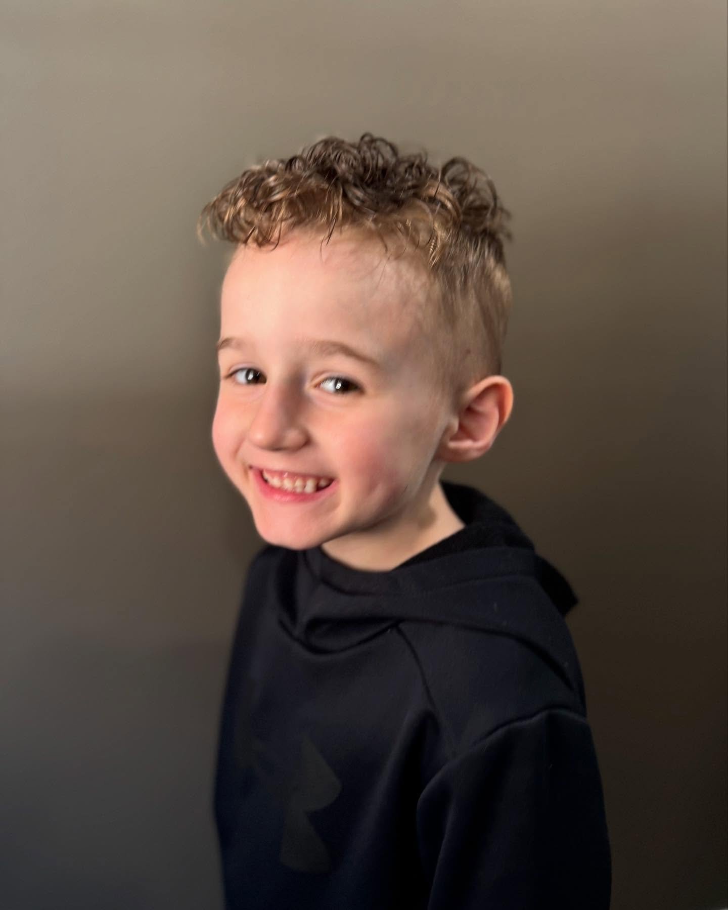 Smiling young boy with curly hair and a black hoodie standing against a gray wall.