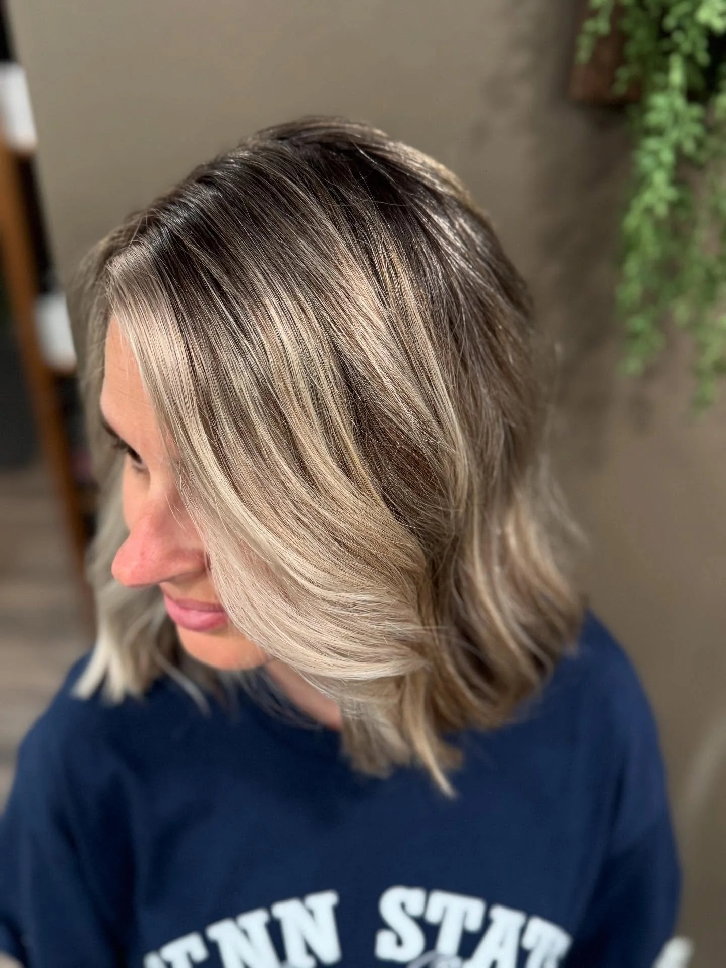 Woman with shoulder-length wavy hair featuring blonde highlights and dark roots, wearing a navy shirt, in an indoor setting with blurred background and some green plants.