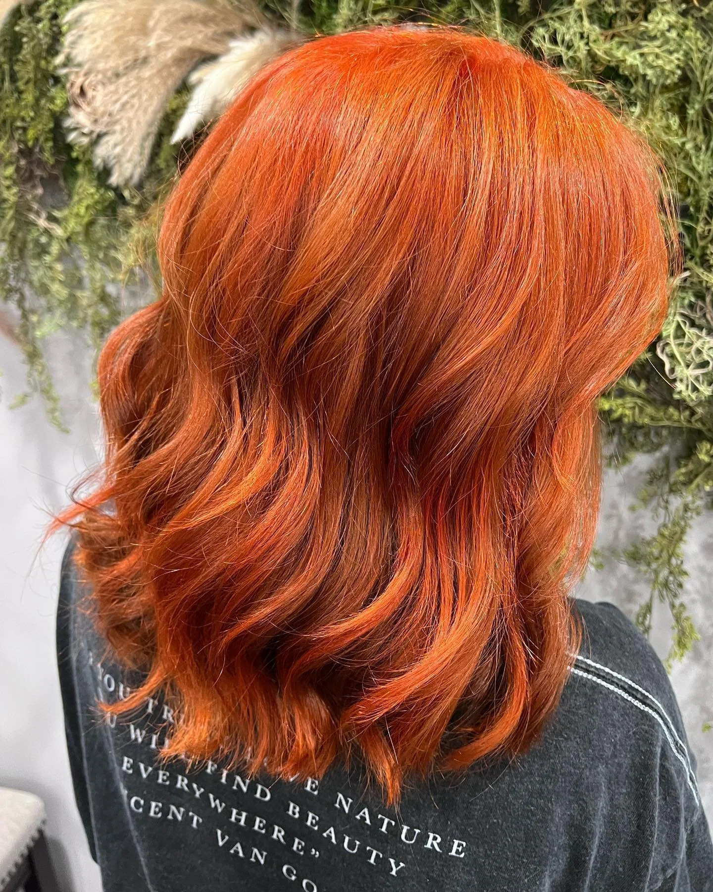 Hair dyed a vibrant orange with soft waves, photographed from the side against a backdrop of green plants.