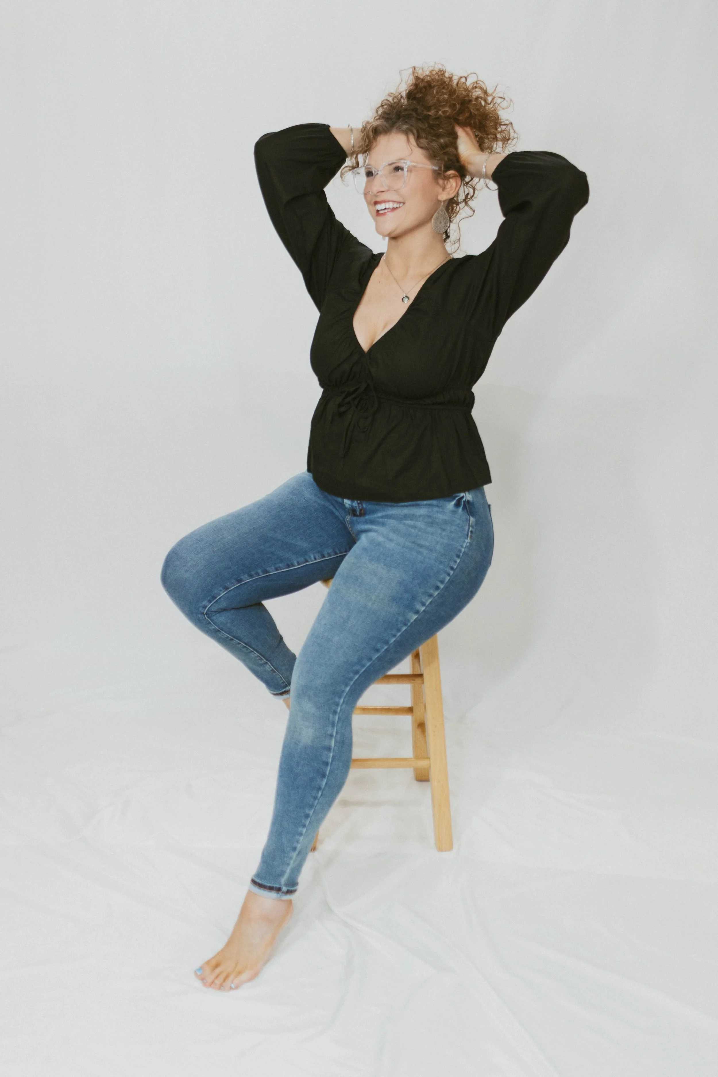 A woman with curly hair and glasses, smiling, sitting on a wooden stool against a plain white background, wearing a black top and blue jeans.