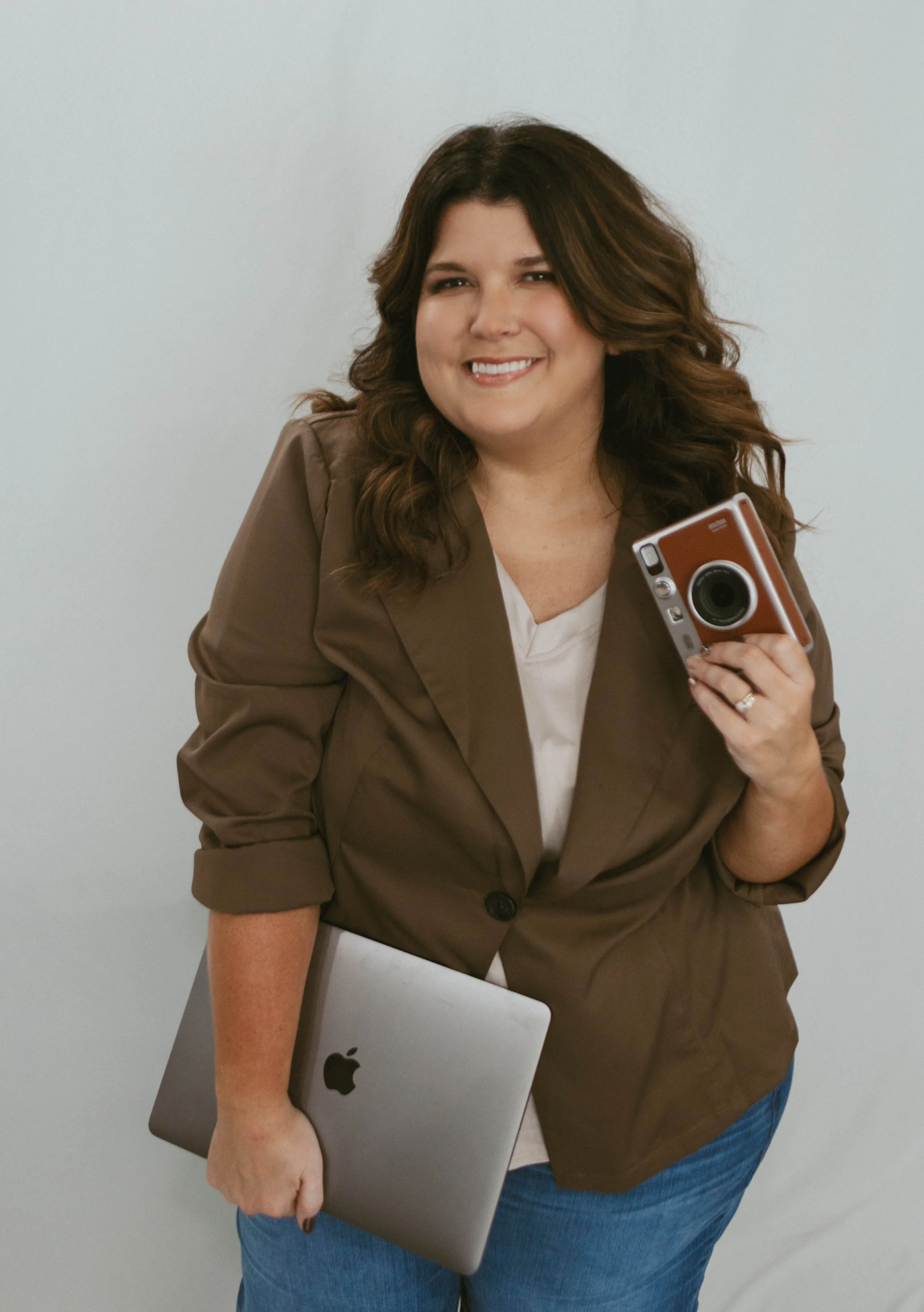 A woman with long brown hair, smiling and holding a camera in her right hand and a closed MacBook in her left hand, standing against a plain light gray wall.