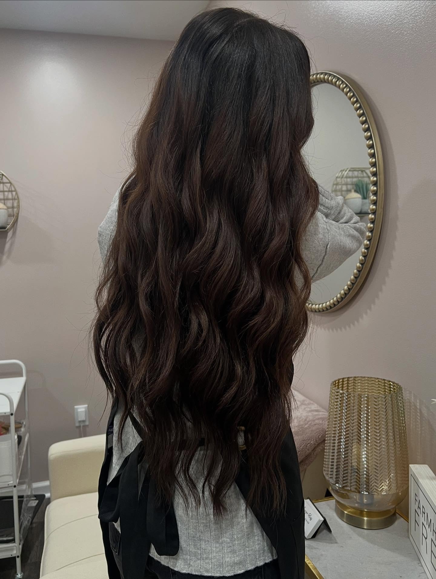 A woman with long, wavy dark brown hair is looking in a mirror with a beaded border, in a room with neutral-colored walls and decorative items.