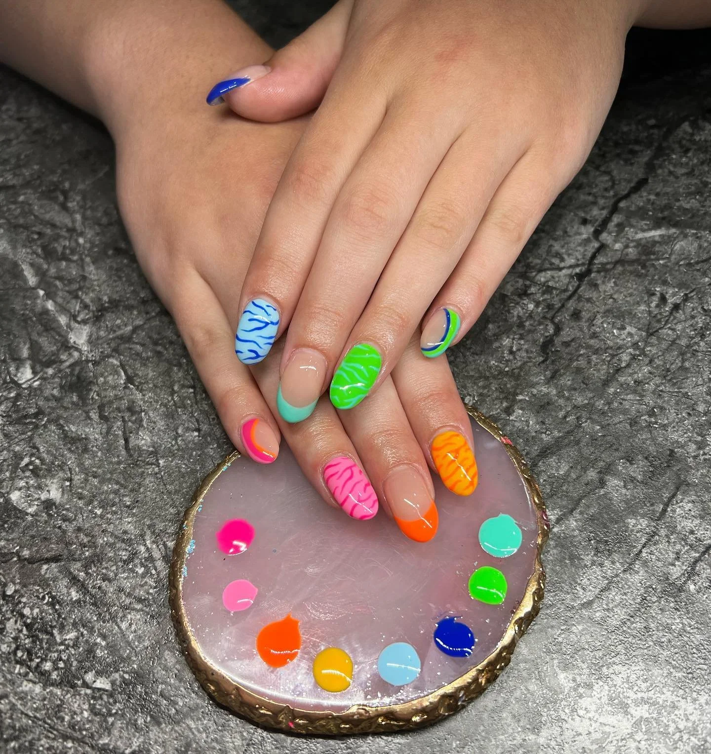 A person's hands with colorful, patterned nail art resting on a pink and gold-bordered circular nail polish palette. The nails feature various bright colors and zebra stripe patterns.