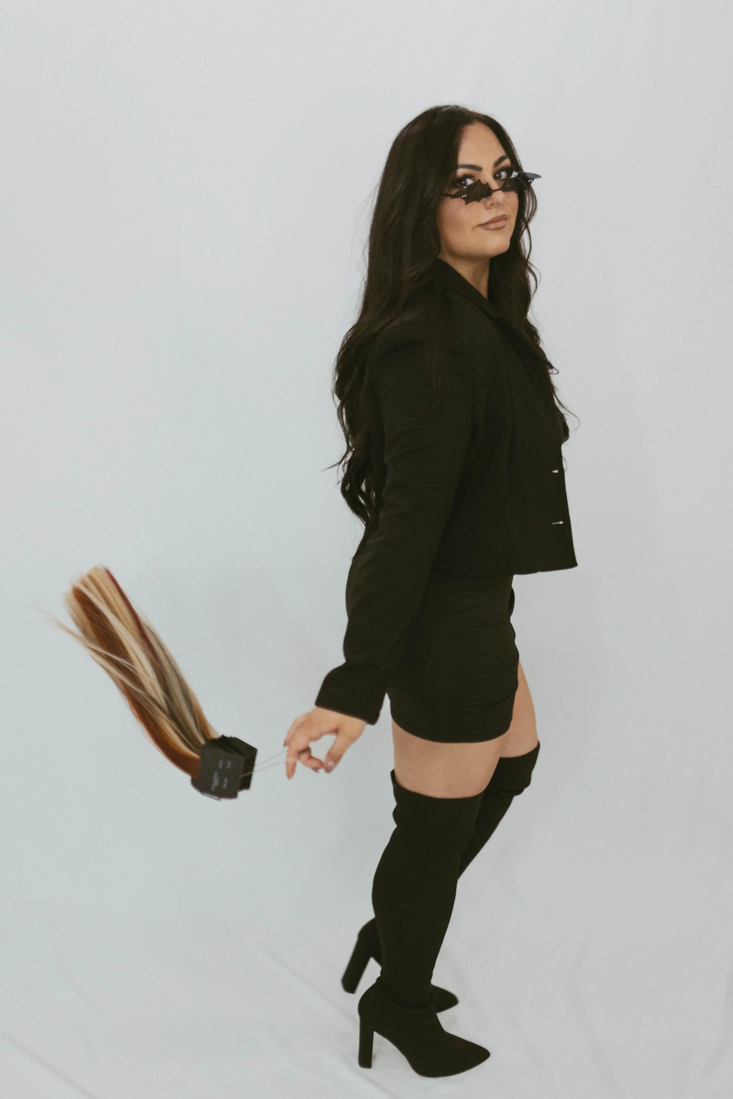 Woman with long dark hair wearing black sunglasses, black blazer, black shorts, and thigh-high black boots, holding a hair extension tester.