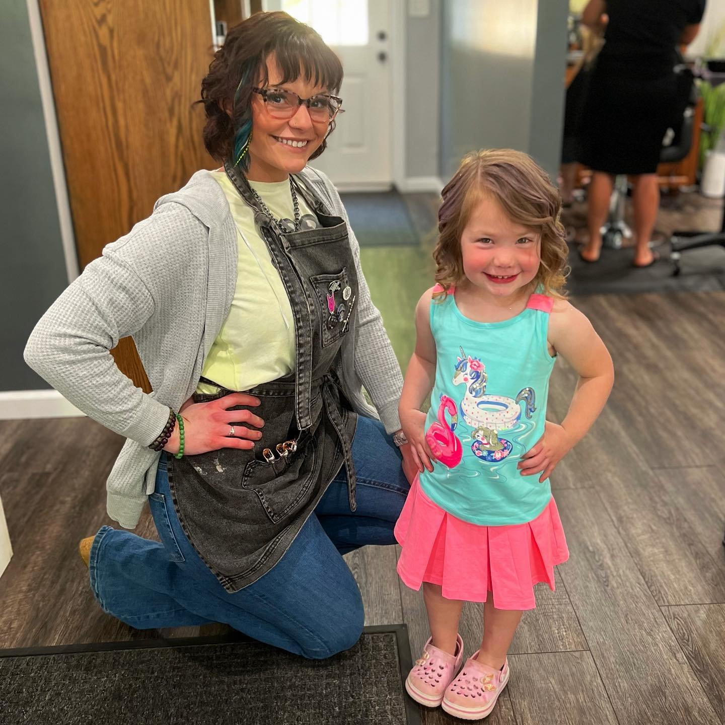 A woman and a young girl posing indoors, both smiling. The woman is kneeling on one knee, wearing glasses, a gray hoodie, and denim jeans. The girl is standing with hands on hips, wearing a turquoise shirt with a unicorn and water design and a pink s