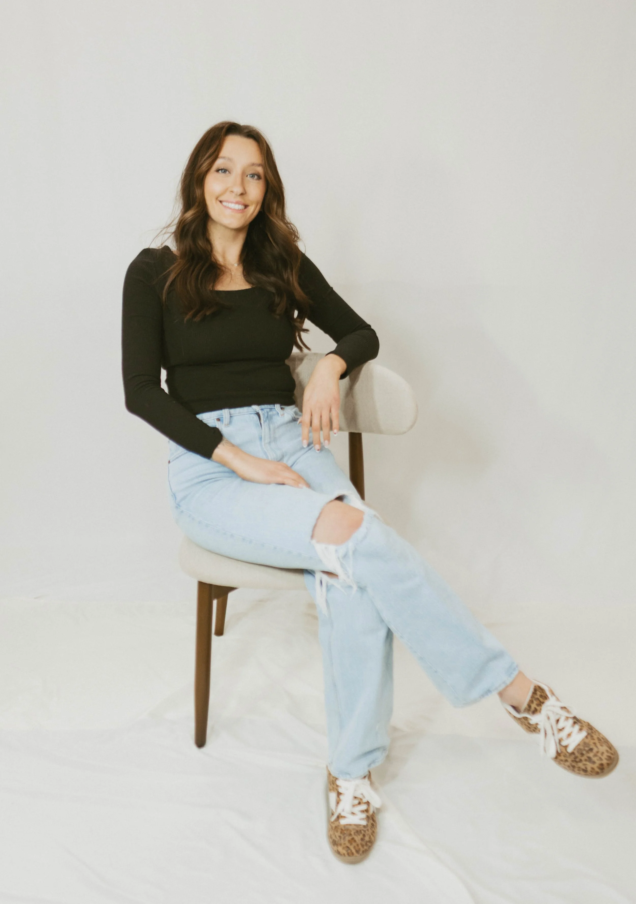 A woman with long wavy hair, glasses, and a black shirt, lying on her stomach on a white surface, smiling and looking at the camera, wearing light blue jeans and leopard print sneakers.