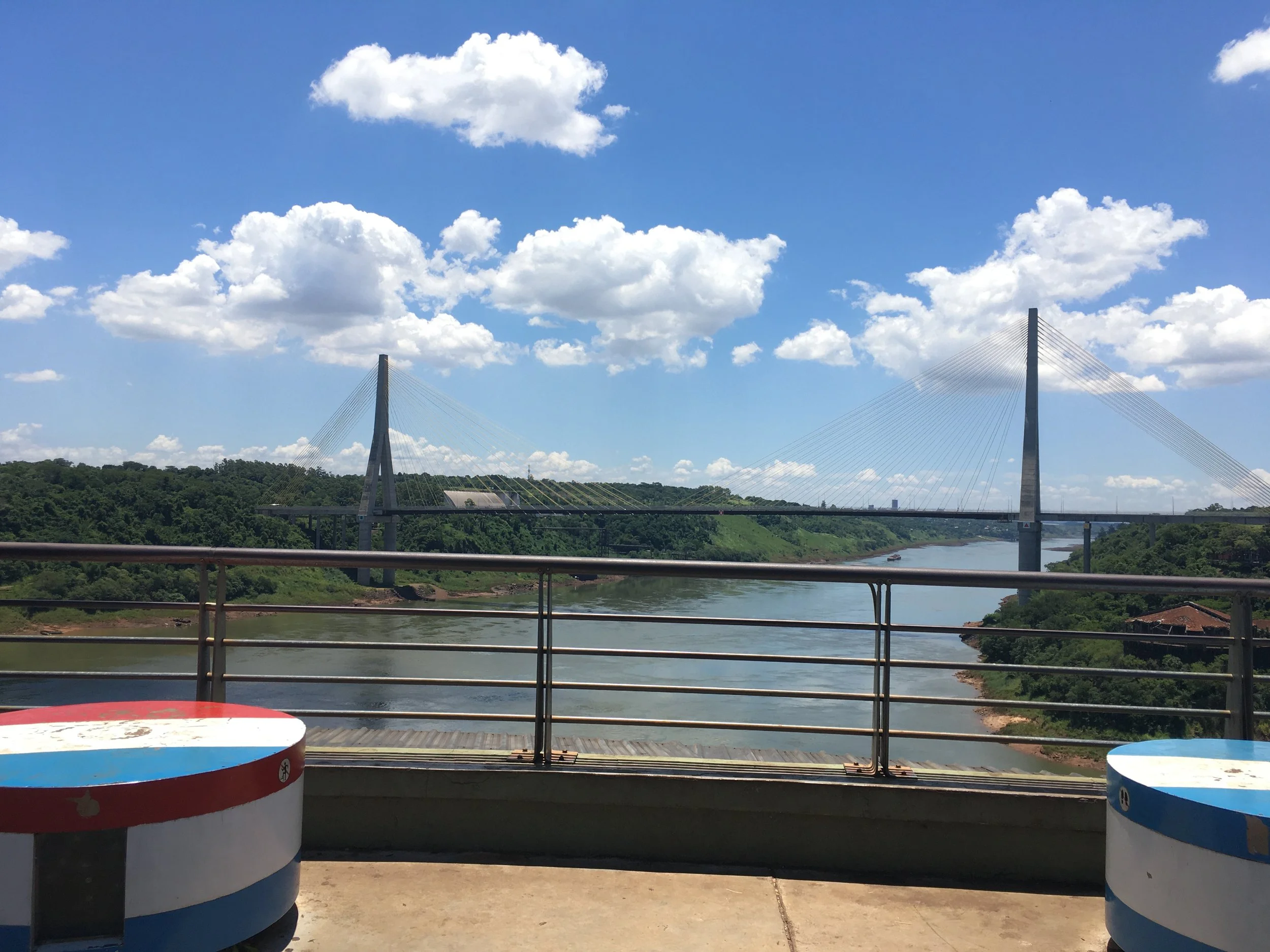 View of a river with a bridge spanning across it, surrounded by green hills under a partly cloudy blue sky, with a railing in the foreground and two circular stools in red and blue.