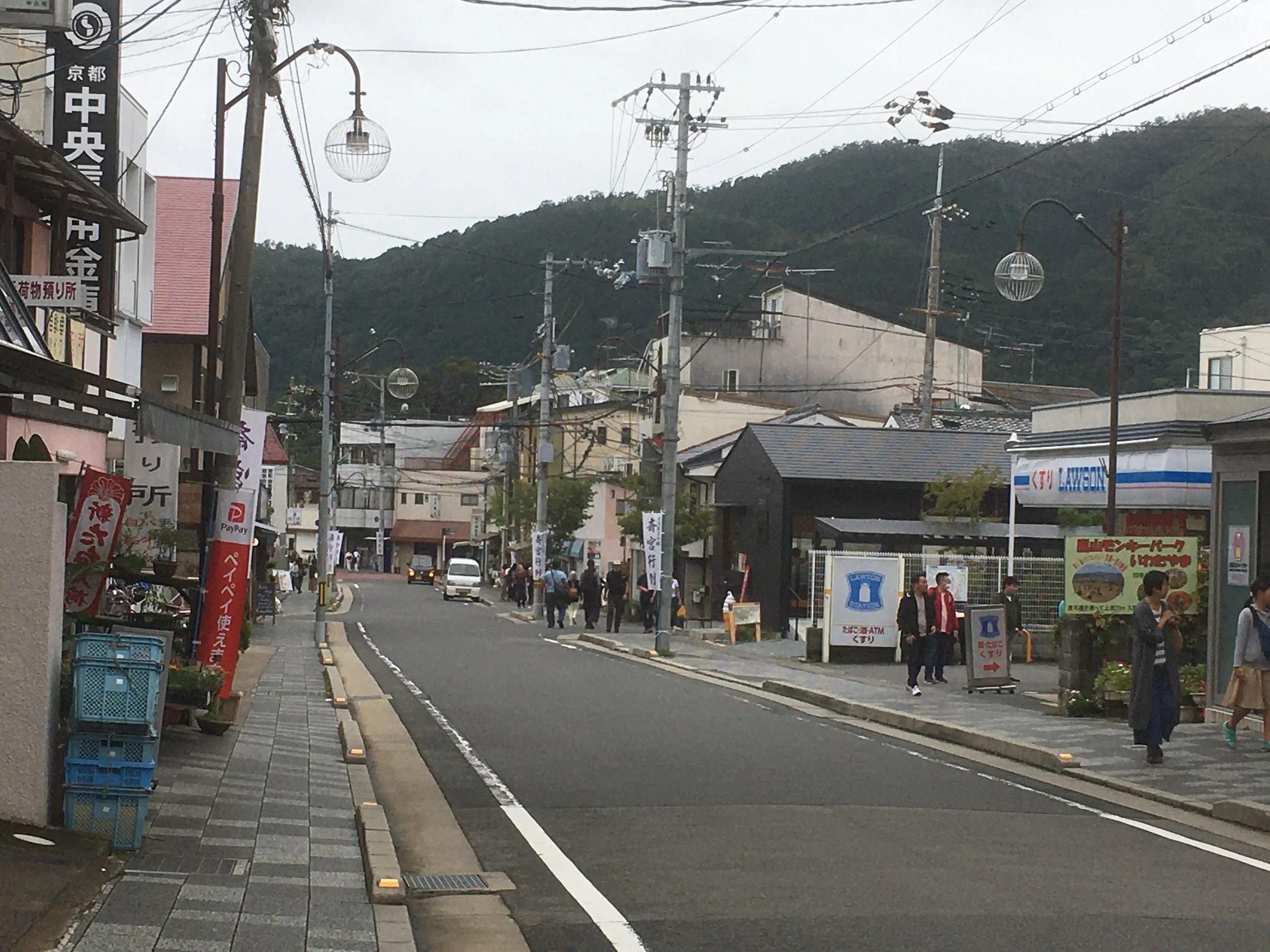 Street view of a small town in Japan with various businesses, pedestrians, cars, and power lines, with a mountain in the background.