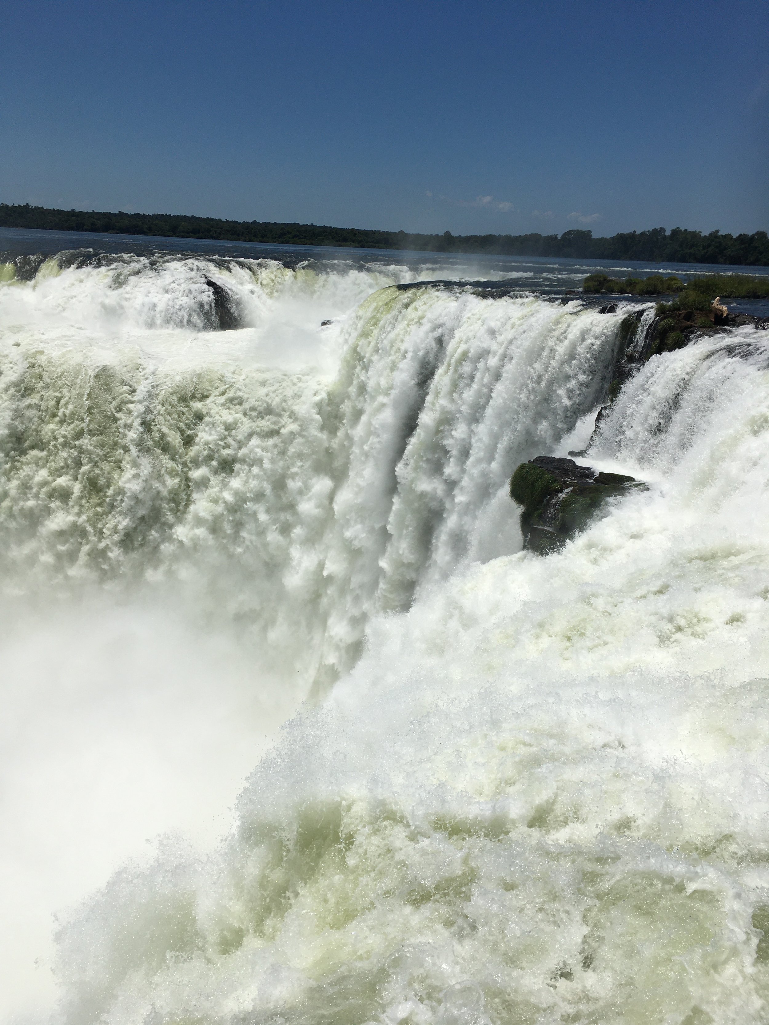 Waterfall cascading over rocks with turbulent white water, lush greenery in the background, and a clear blue sky.