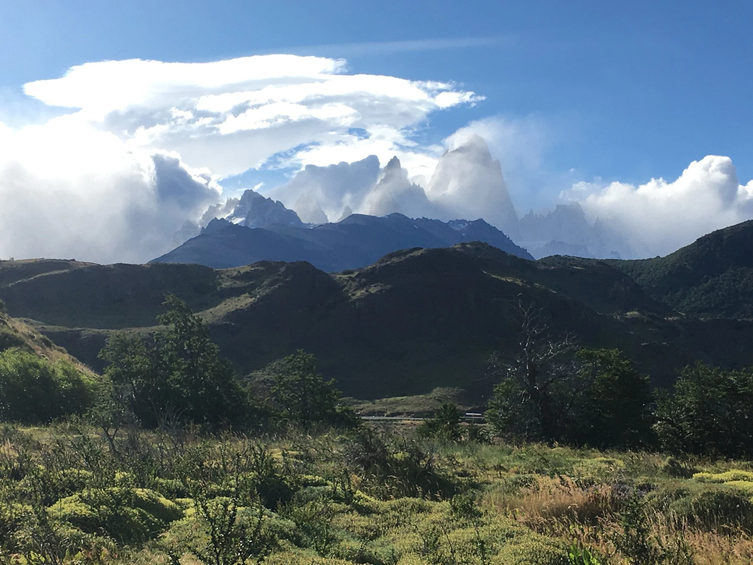 Scenic view of green hills and mountains with clouds and snow-capped peaks in the background.