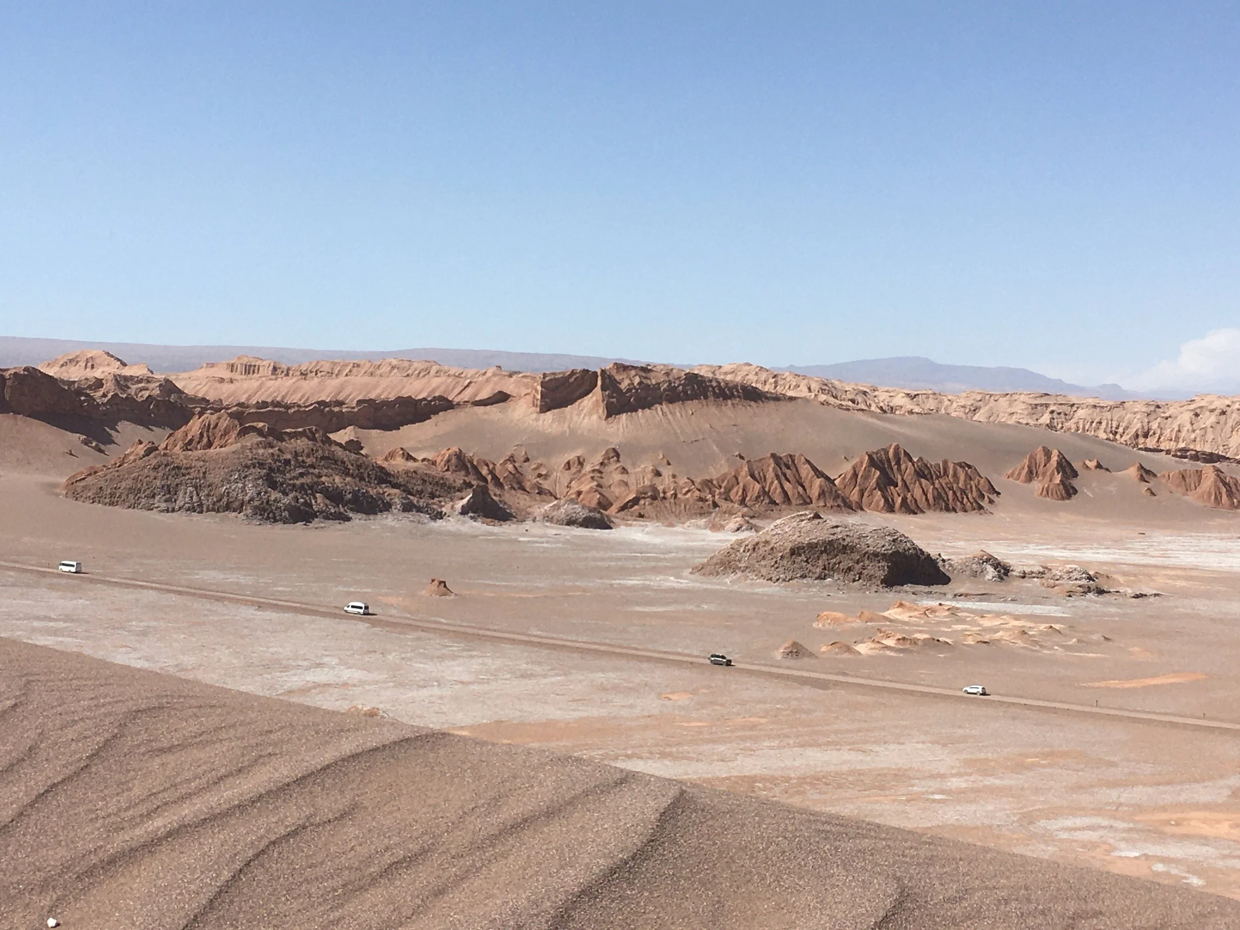 Desert landscape with rocky mountains, sandy dunes, and a clear blue sky. Four vehicles are driving along a dirt road.