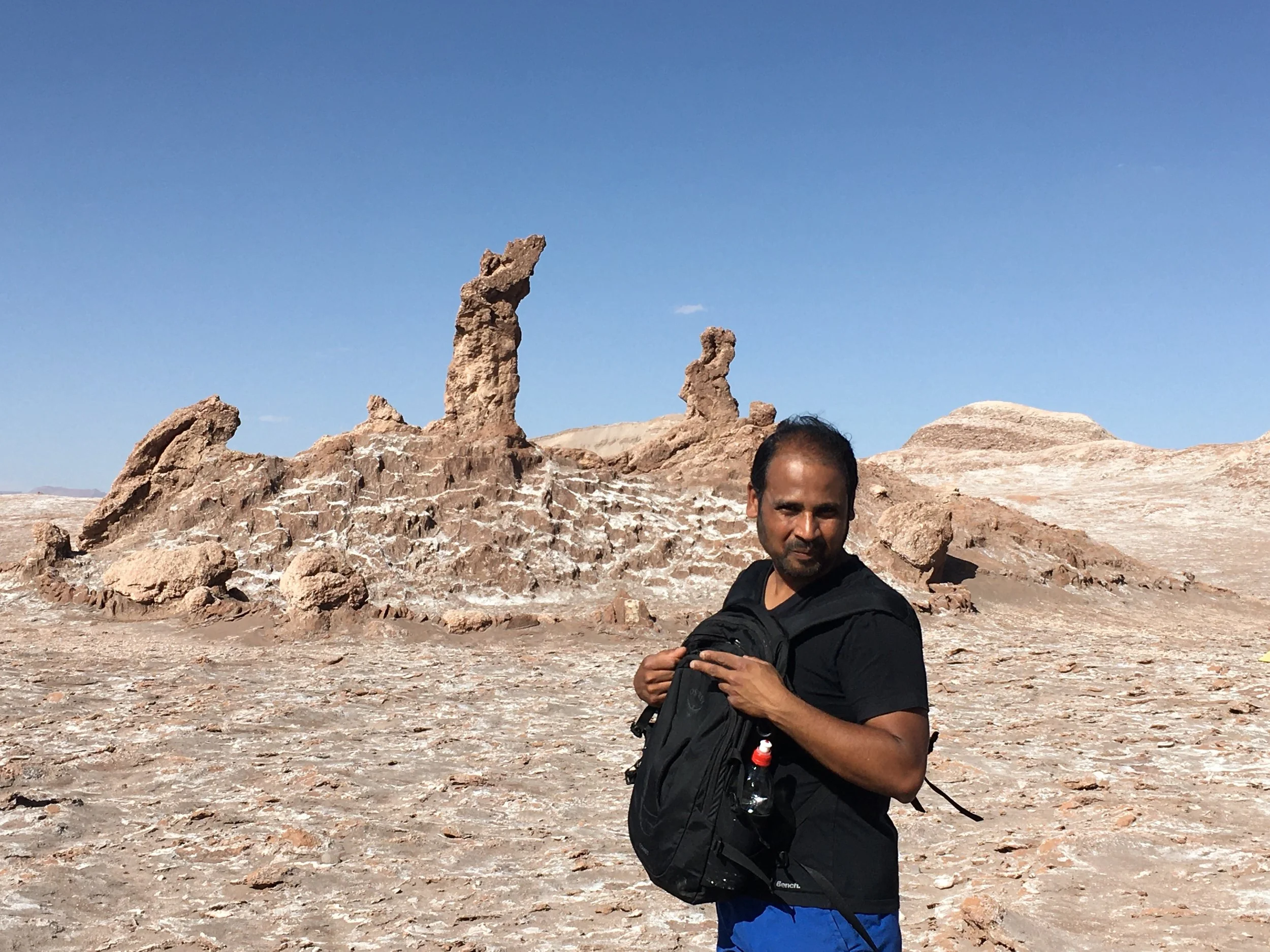 A man with dark hair, wearing a black shirt and blue shorts, standing in a desert with rock formations in the background under a clear blue sky.