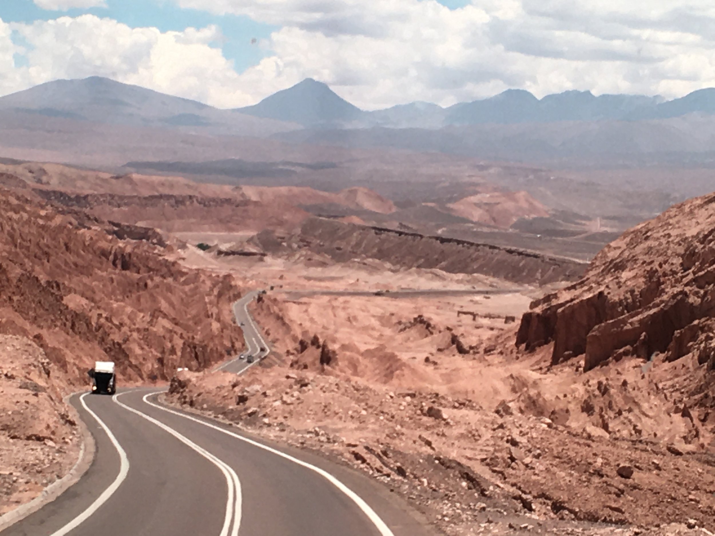 Winding road through a desert landscape with reddish-brown rocks and mountains in the distance under a cloudy sky.