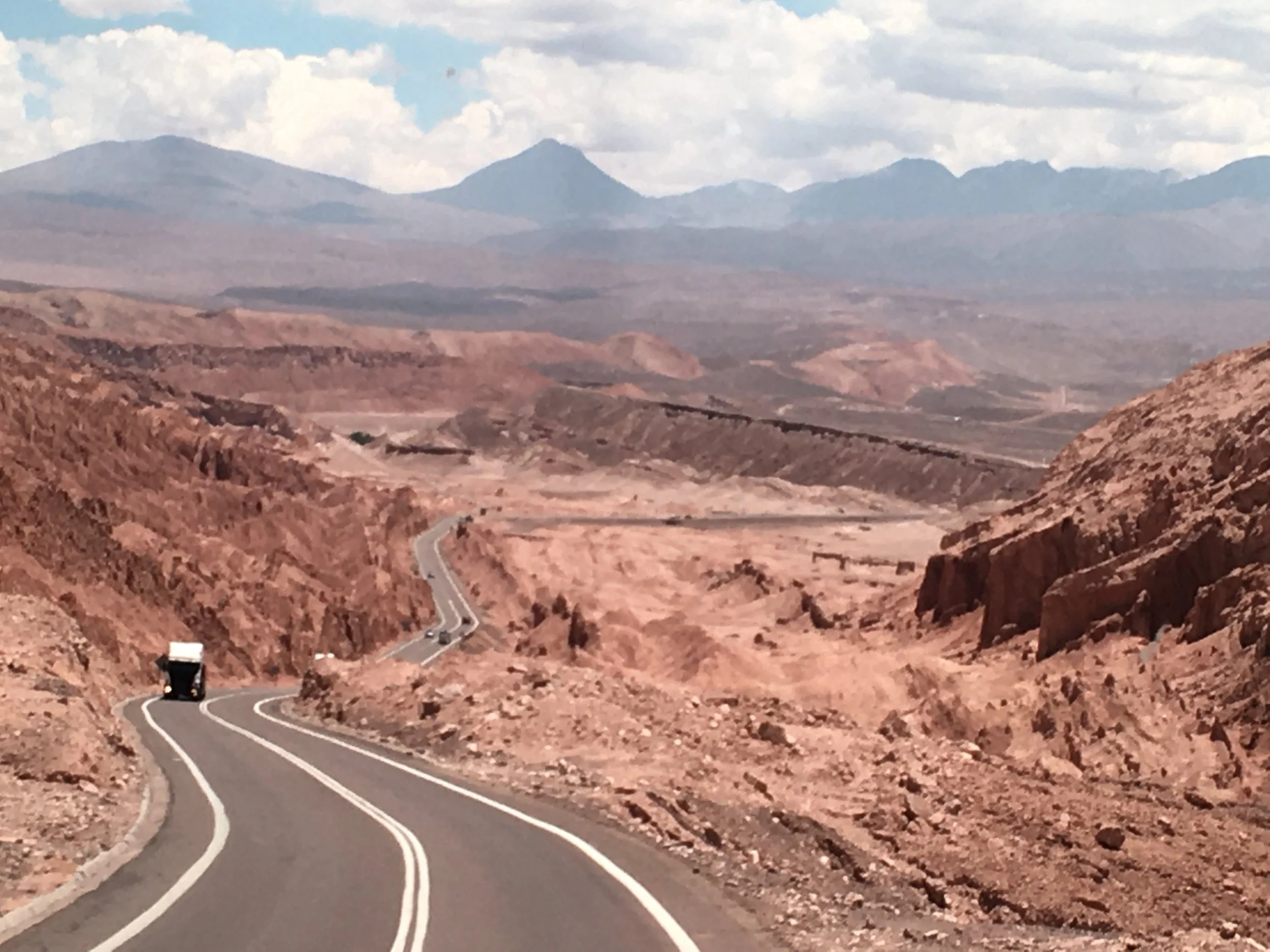 A winding road through a desert landscape with reddish-brown terrain and distant mountains under a partly cloudy sky.