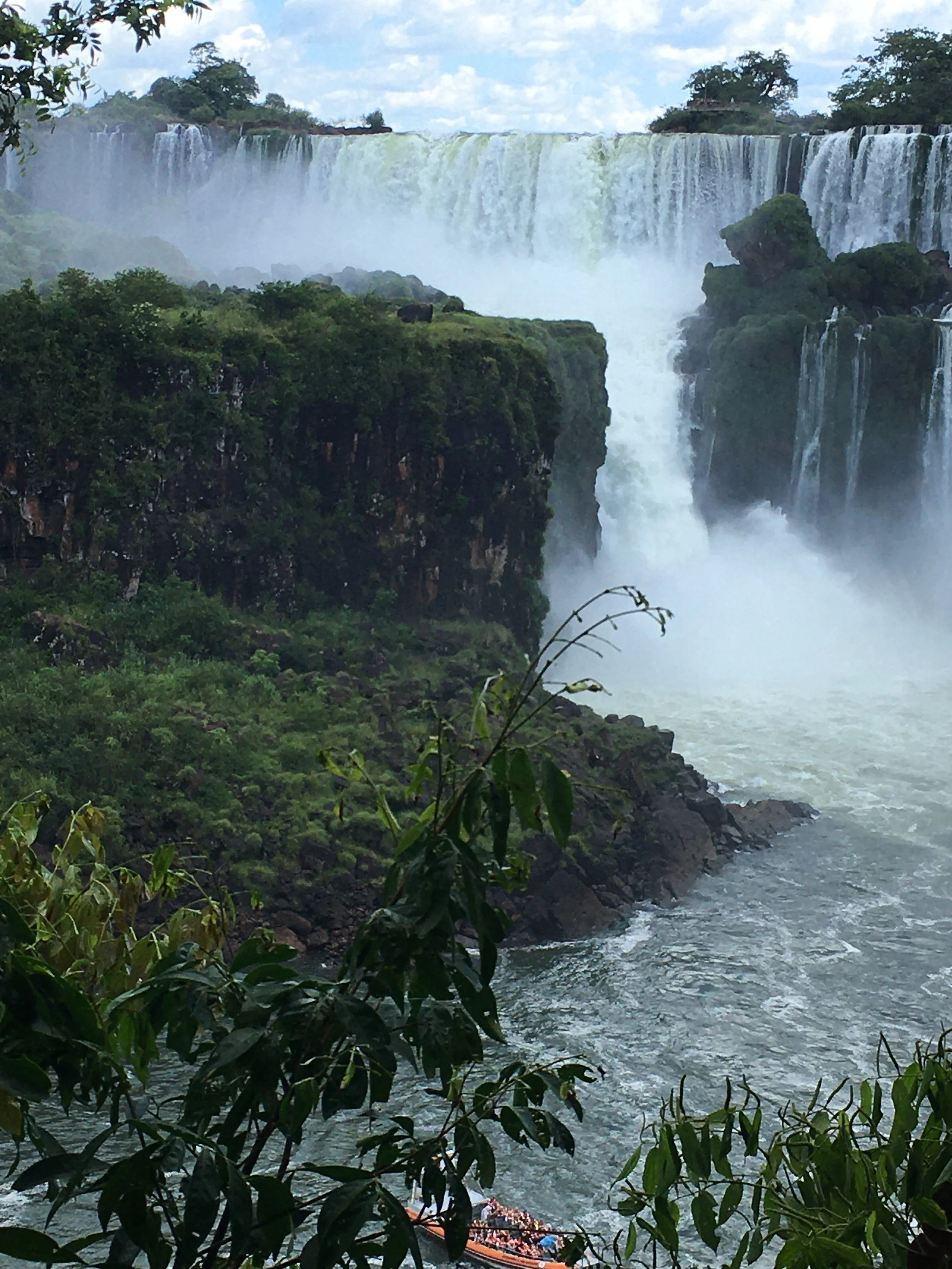 Niagara Falls with cascading waterfalls, lush green cliffs, mist rising from the water, and a boat near the base of the falls.