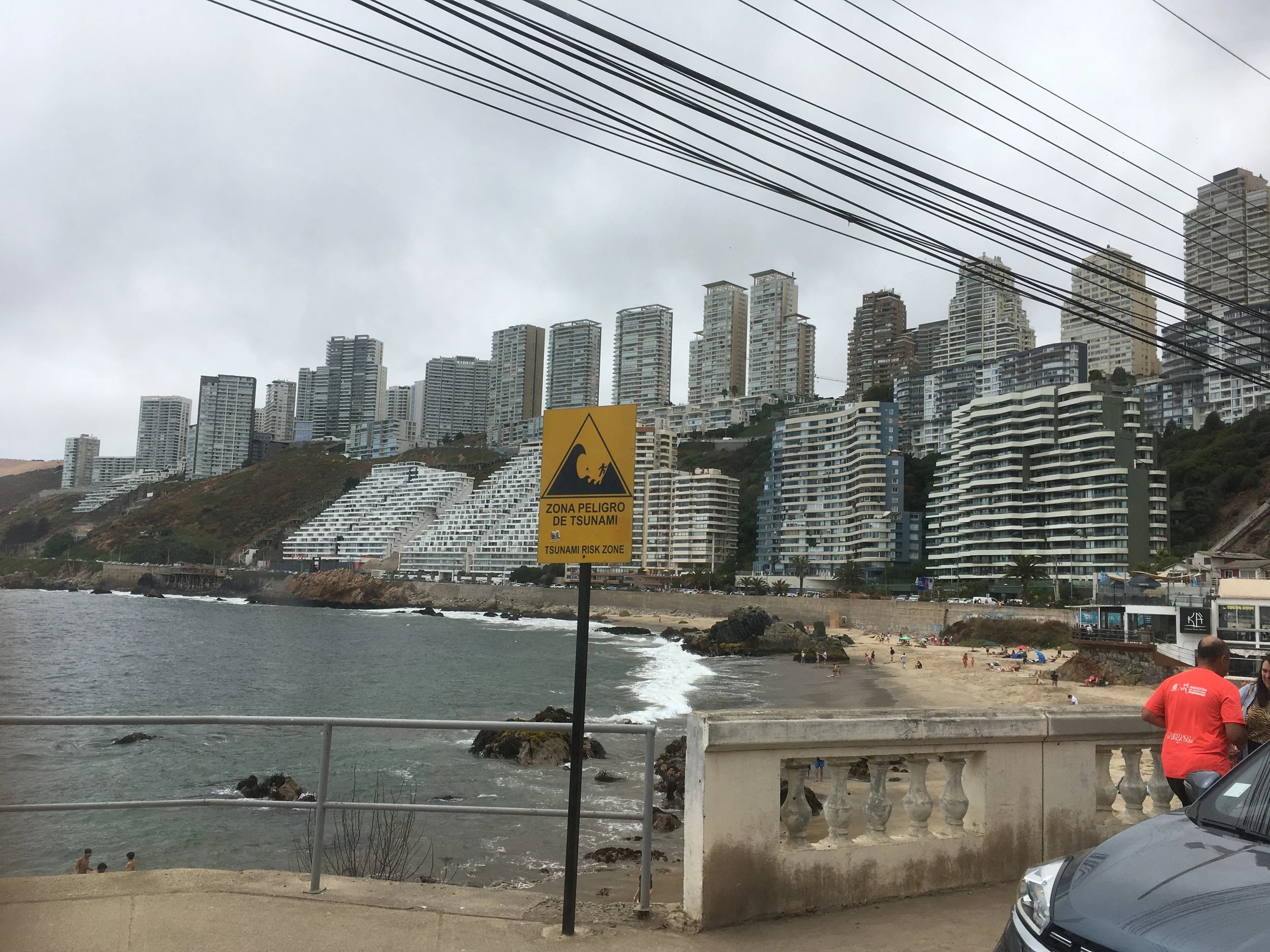 Beach with people swimming, a warning sign indicating a tsunami risk zone, and a backdrop of high-rise buildings on a cloudy day.