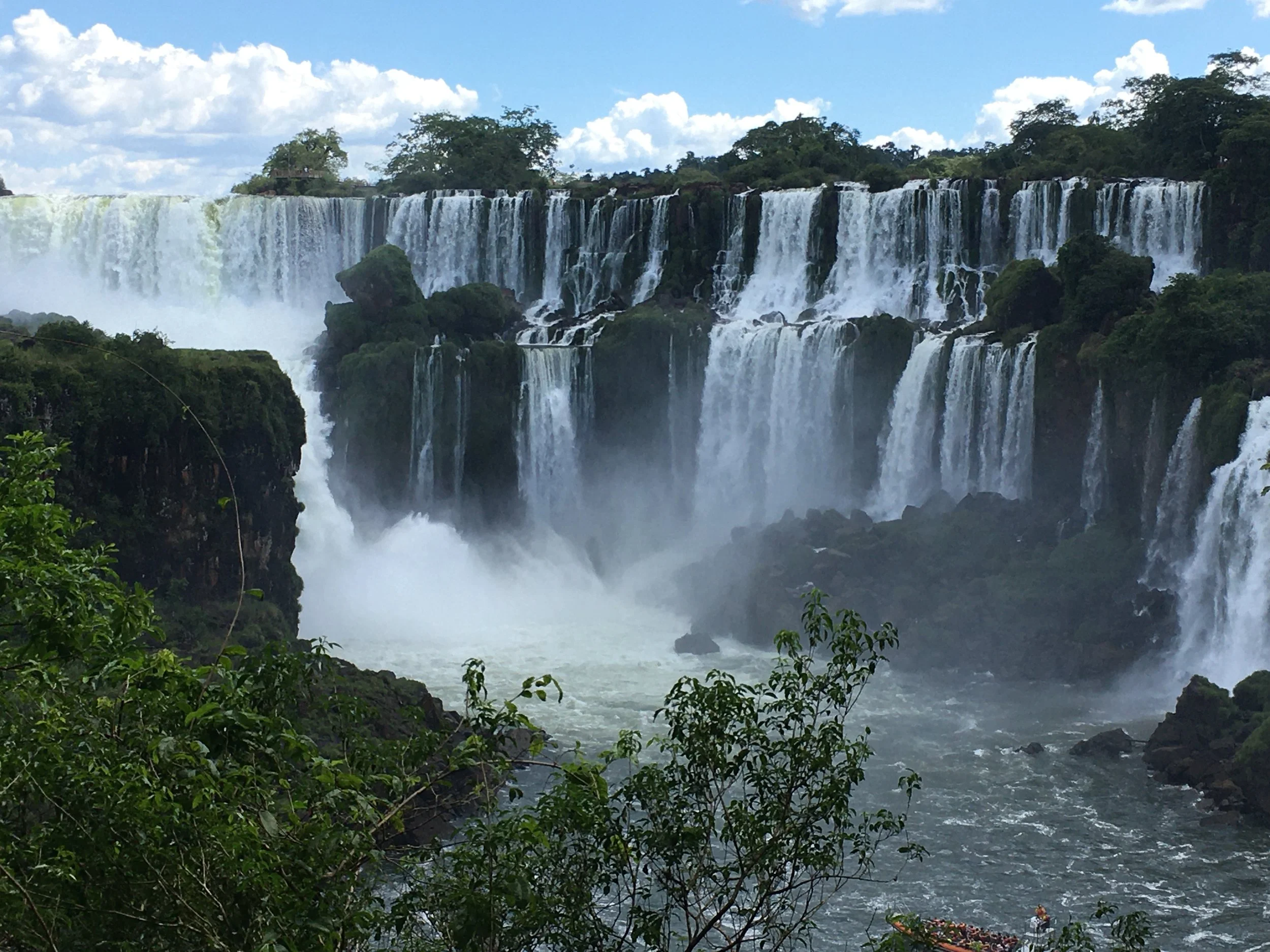 Waterfalls cascading over a cliff with lush greenery in the foreground and trees against a partly cloudy sky.