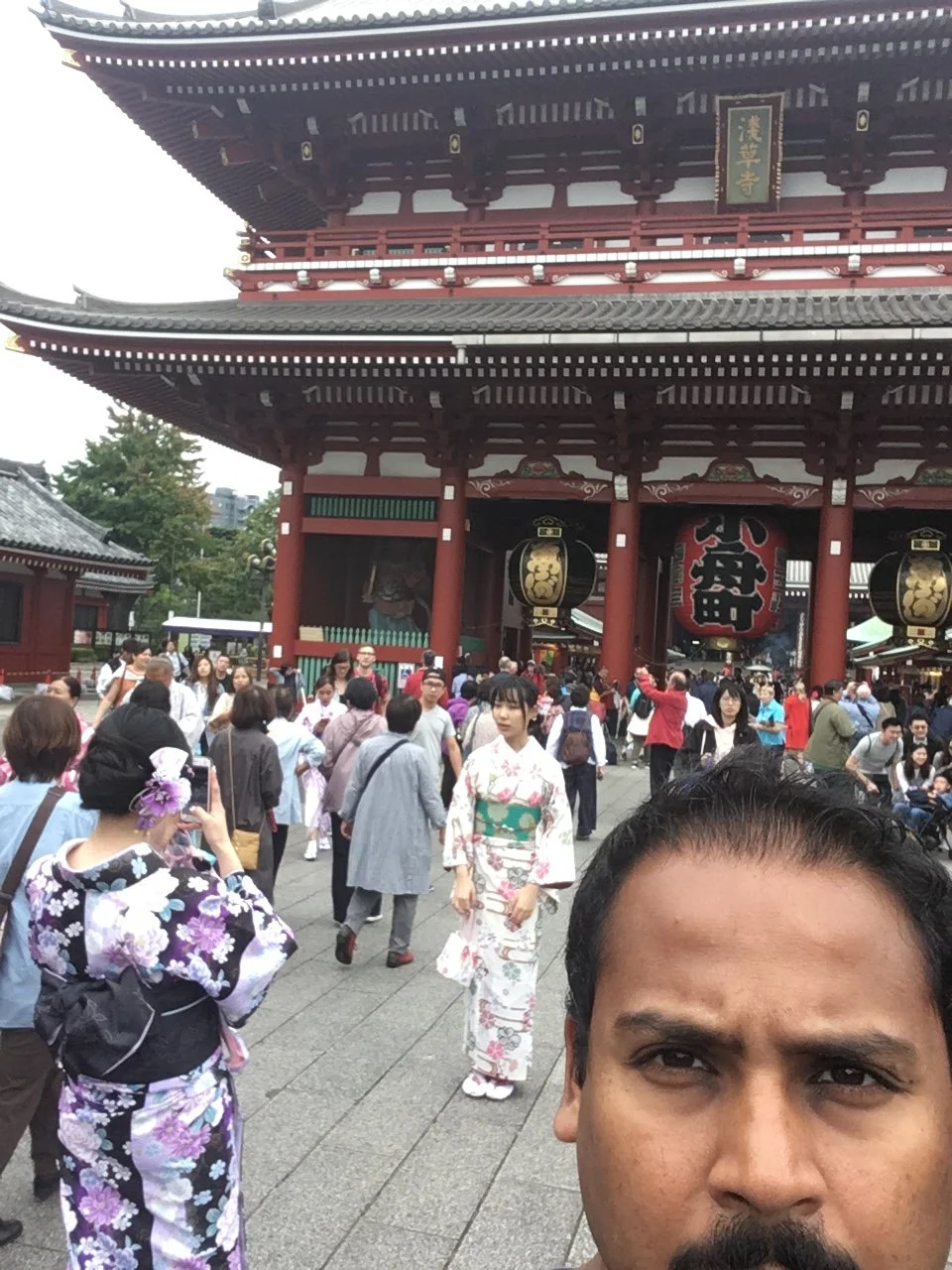 A busy entrance to a traditional Japanese temple with many people, including women in kimonos. The large temple gate features red and black colors with hanging lanterns.