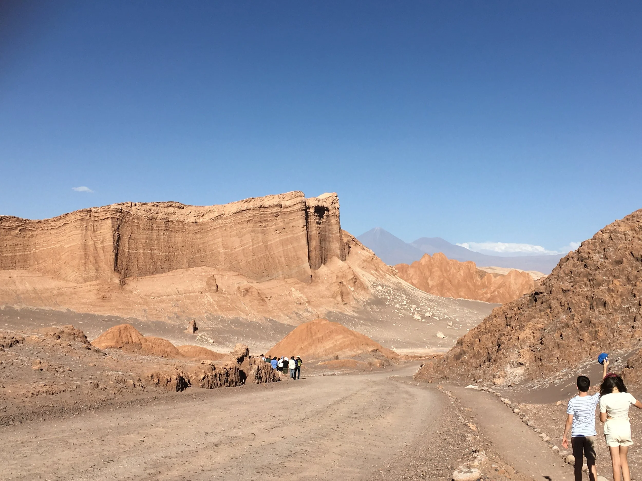 A group of people walking along a dirt path in a desert landscape with large rocky formations and mountains in the background under a clear blue sky.