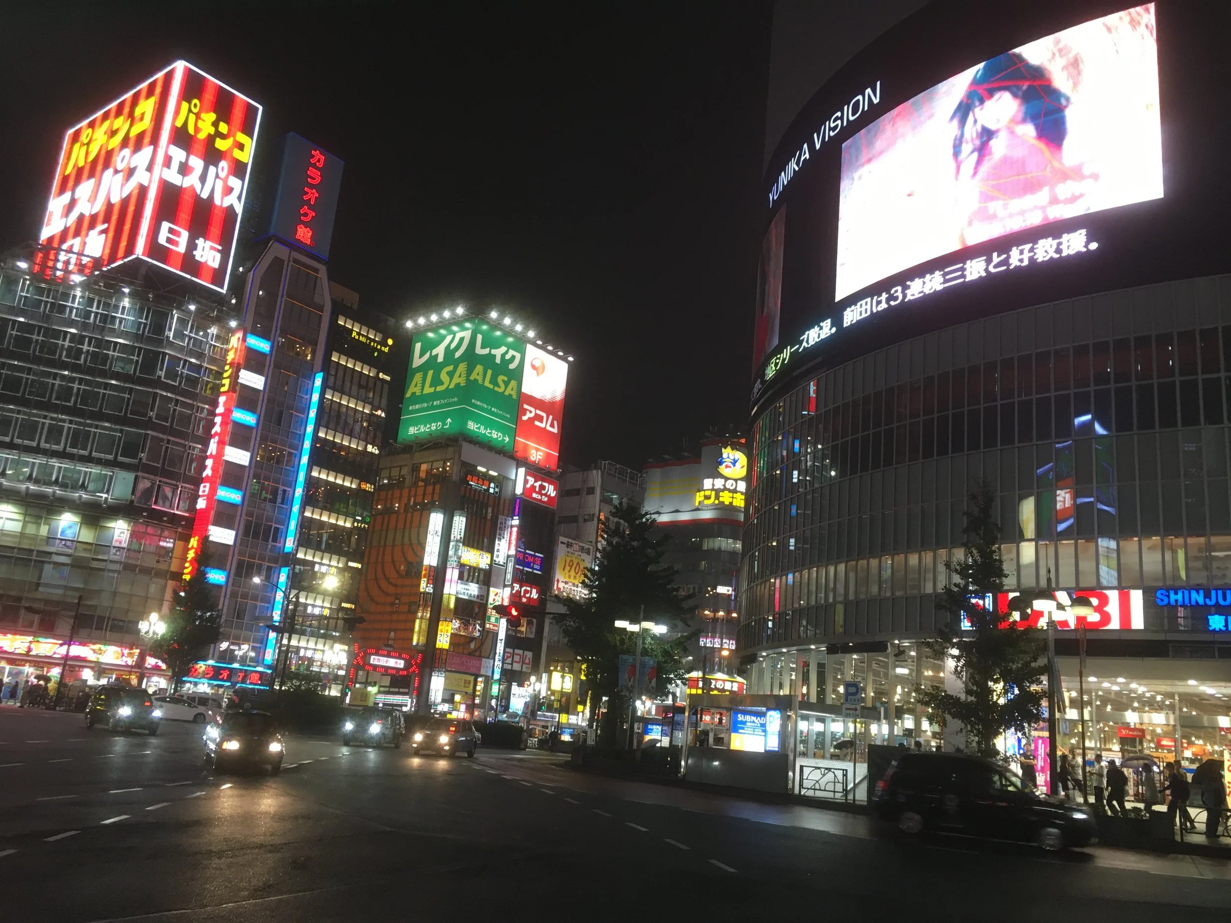 Night view of a busy city street with brightly lit buildings and billboards, including a large curved digital display, in an urban area of Japan.