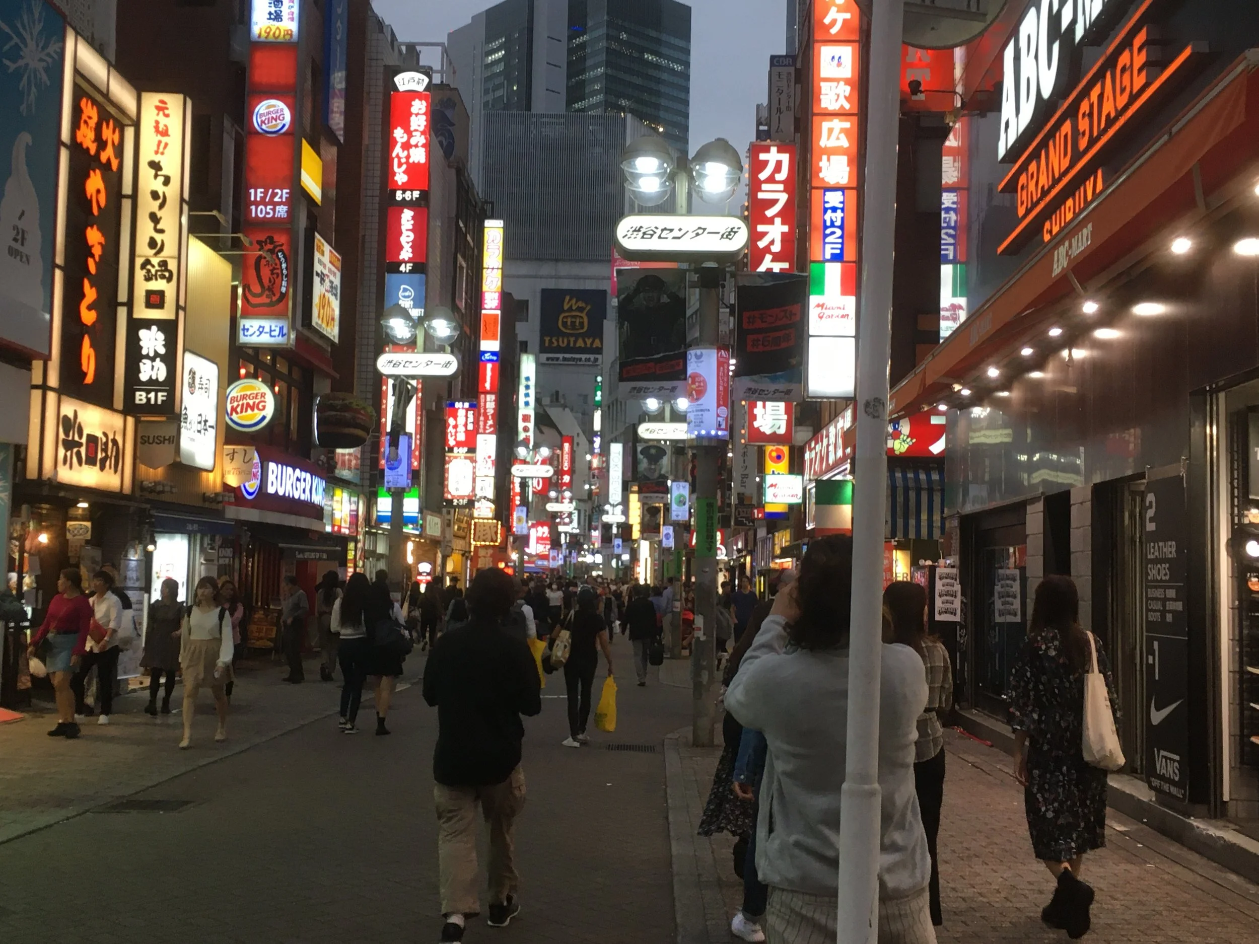 Nighttime busy street scene in an urban area with numerous illuminated signs and advertisements in Japanese. People walk along the sidewalk and street, some looking at signs and taking photos. Bright shop signs include Burger King and other retail ou