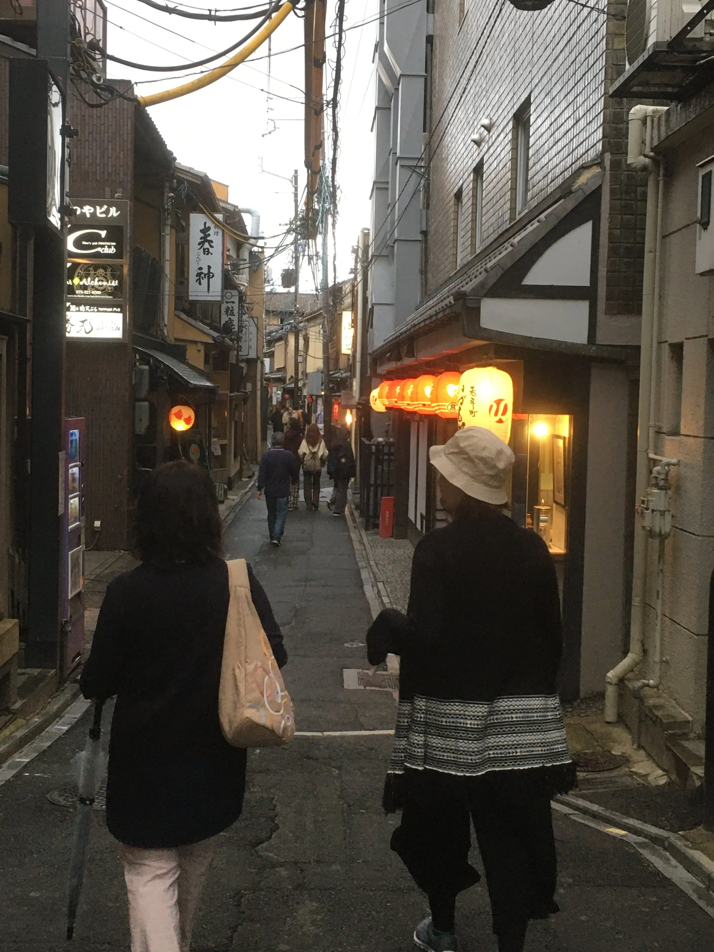 A narrow street in Japan with lanterns hanging outside restaurants, walking pedestrians, and traditional signage.