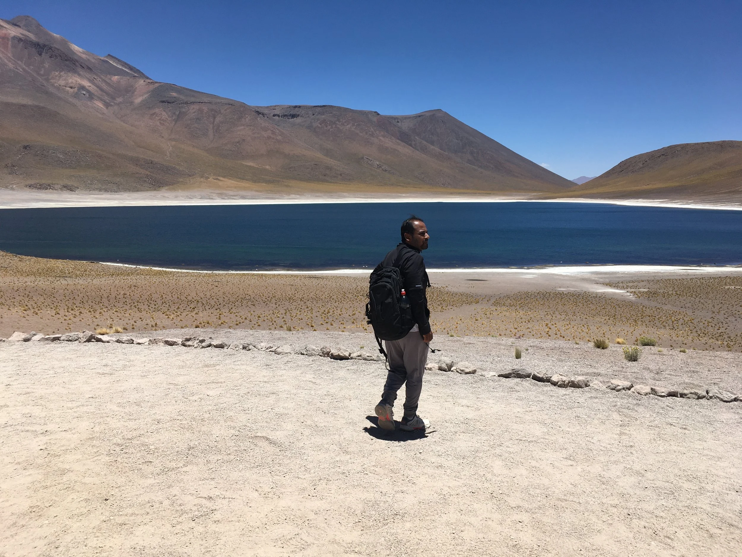 A man hiking with a backpack standing on a sandy trail near a blue lake, surrounded by mountains under a clear blue sky.